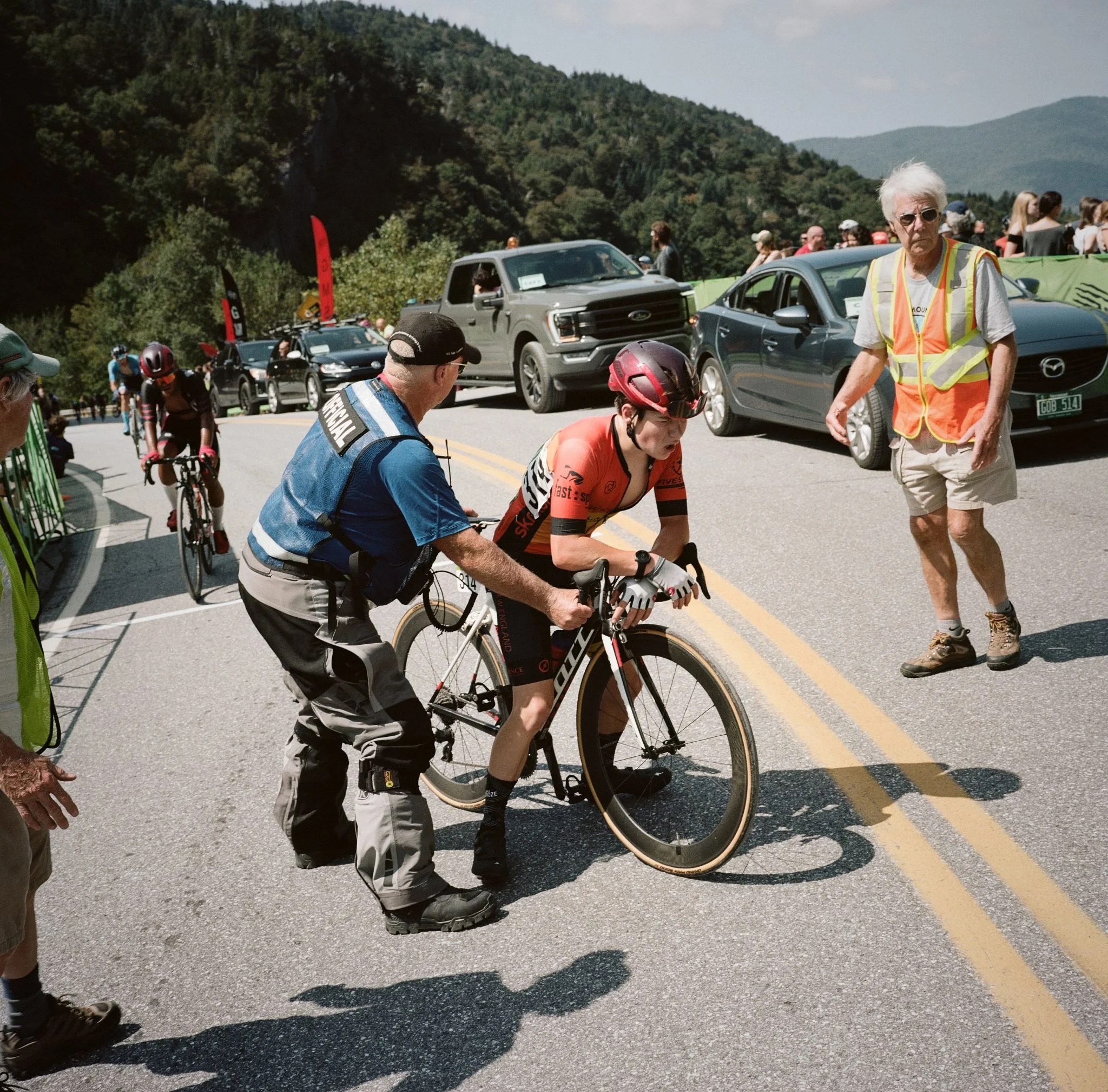 Cyclist in red jersey receives support from staff member during mountain road race with spectators and parked cars lining the route.