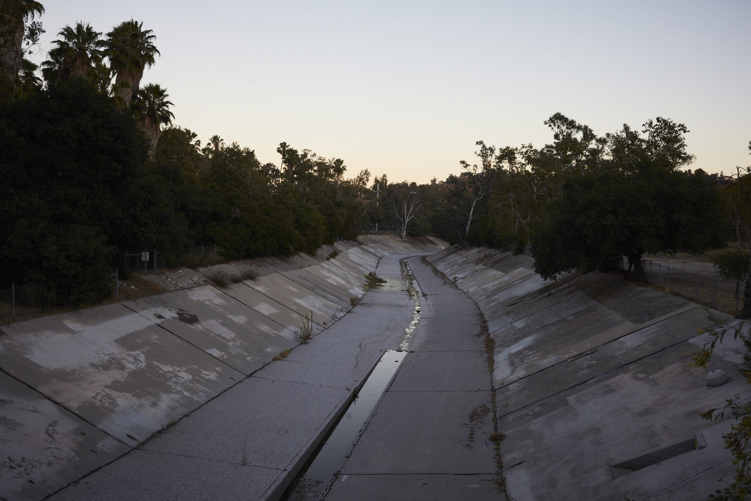 Concrete flood control channel with vegetation and palm trees along the banks at dusk.