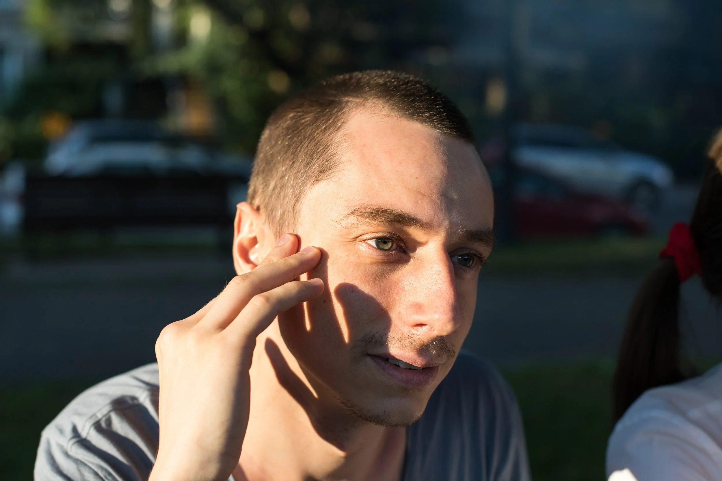 A man in a gray shirt touches his ear while sitting outdoors in afternoon sunlight, with blurred vehicles and greenery in the background.