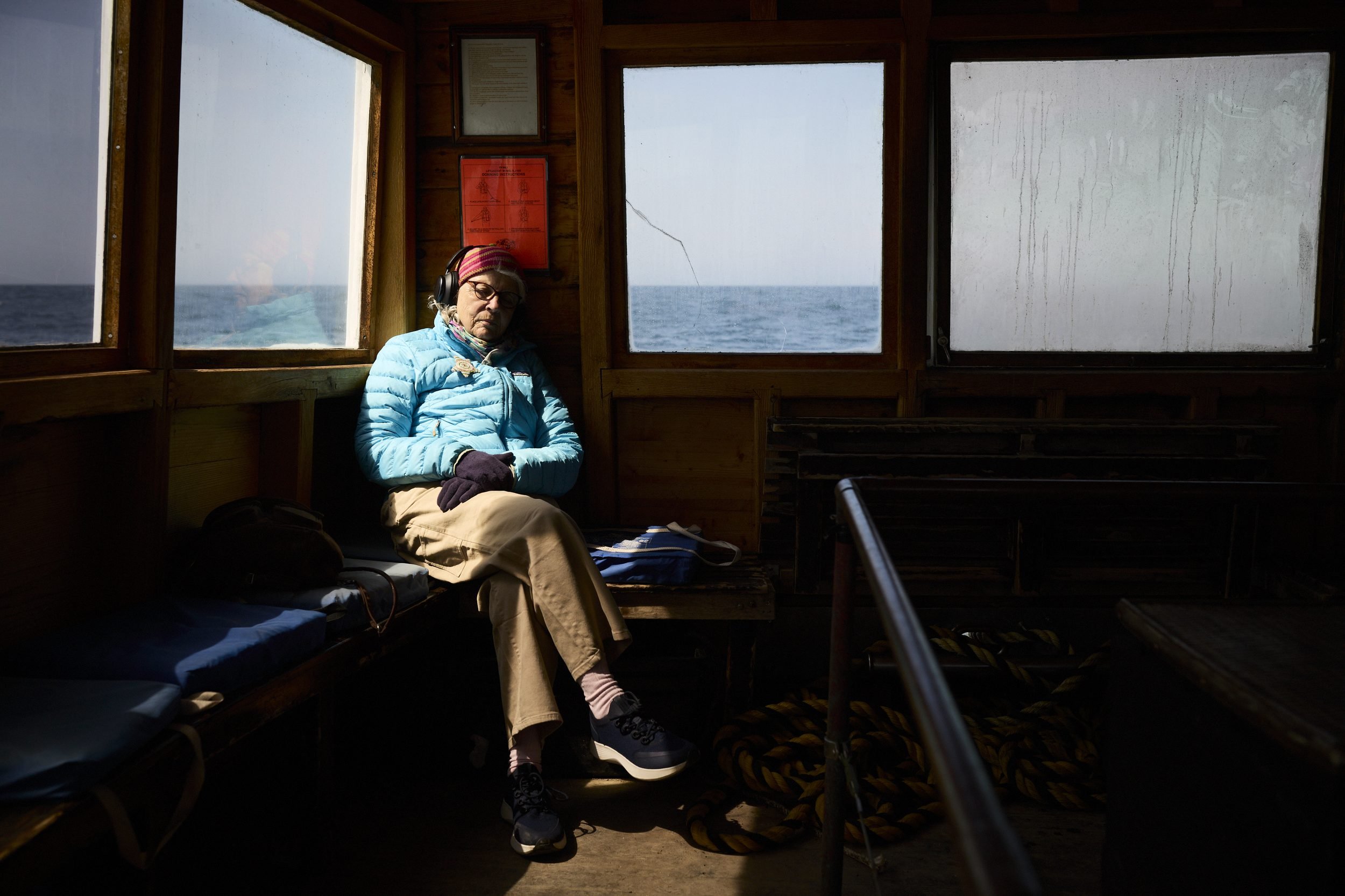 A person sits on a bench inside a boat cabin, gazing out at calm ocean waters through multiple windows.