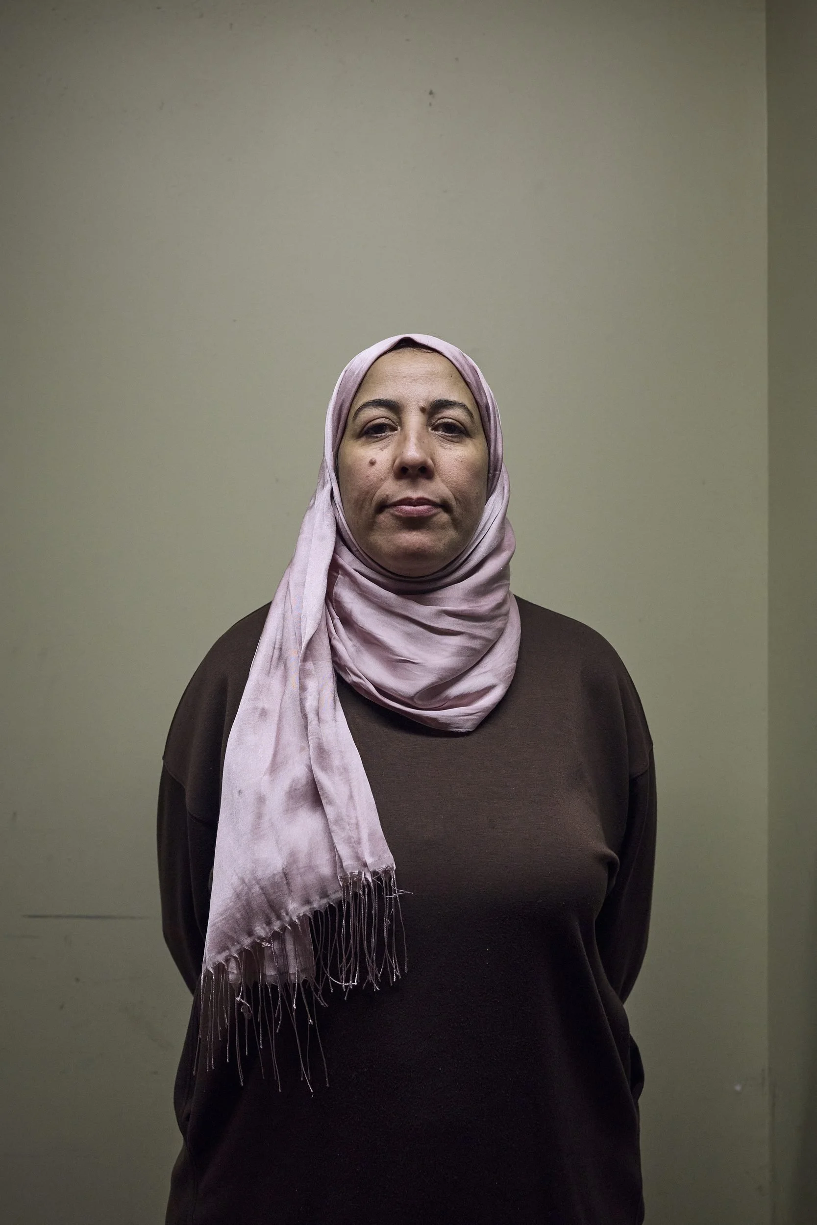 Woman wearing a lavender hijab and dark shirt stands against a plain beige wall.