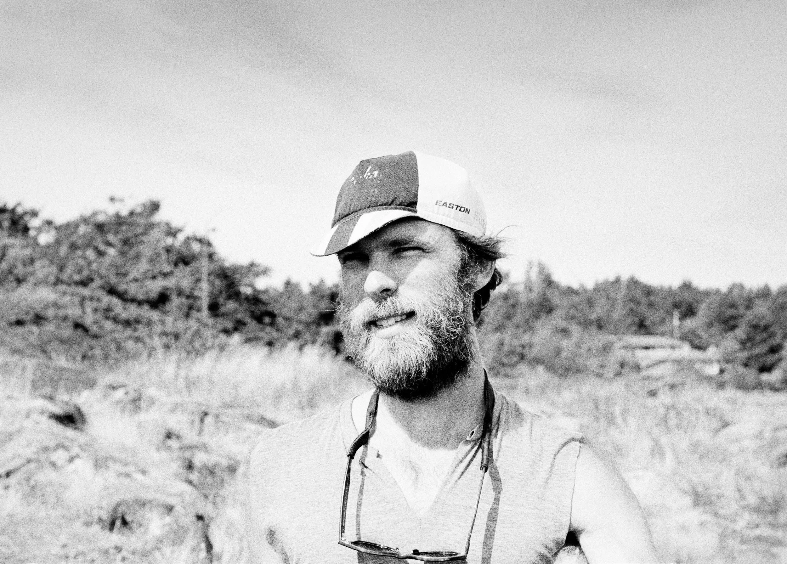 Bearded man wearing a baseball cap and sleeveless shirt stands in a sandy, forested area.