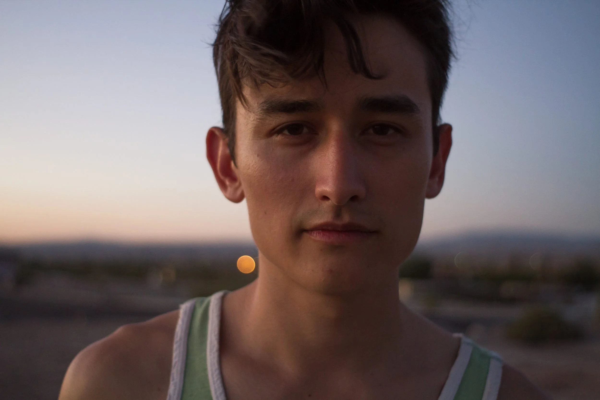 Young man in a tank top looking at camera during golden hour with landscape visible behind him.