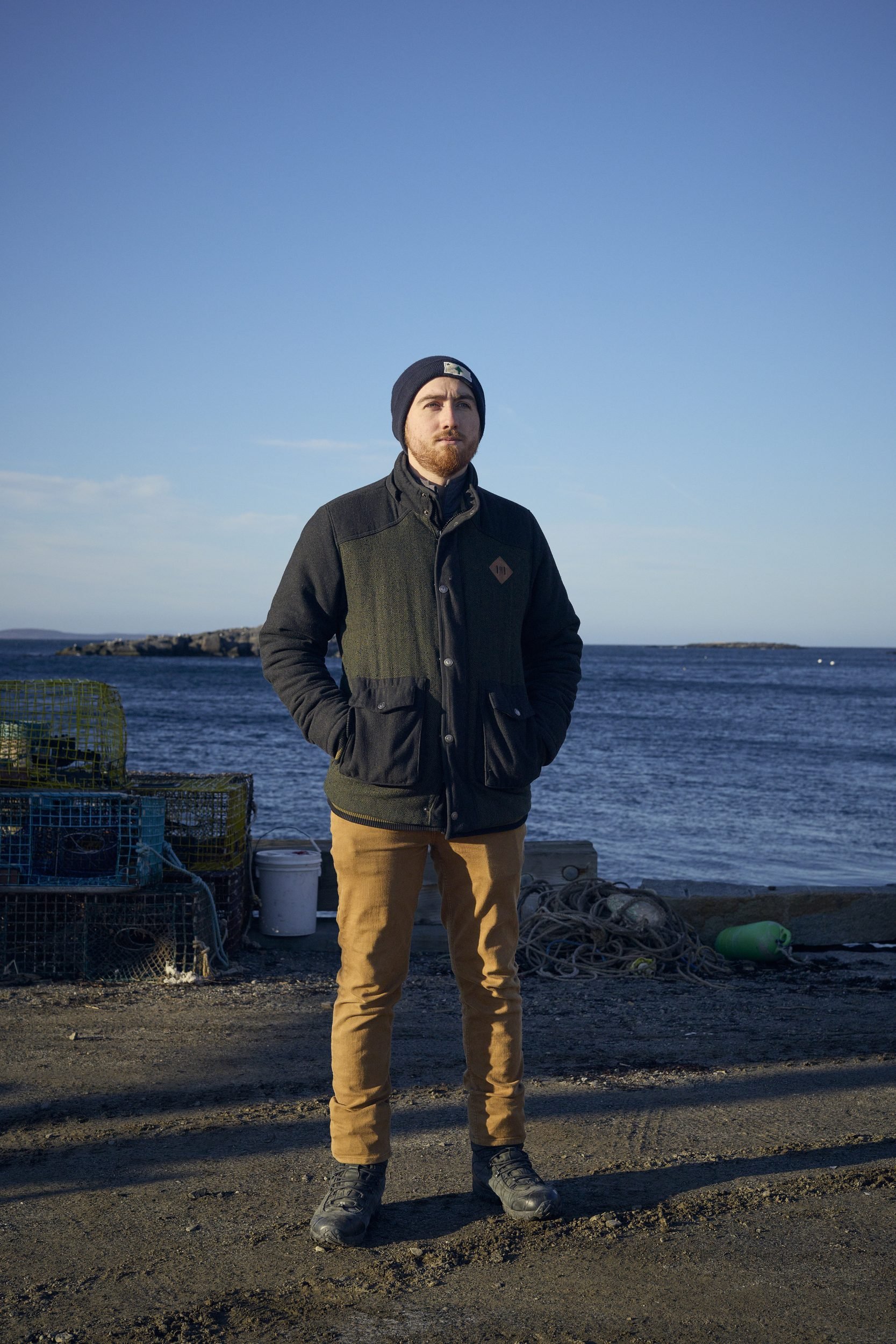 Man in dark jacket and yellow pants stands on a rocky beach with fishing traps and ocean in background.