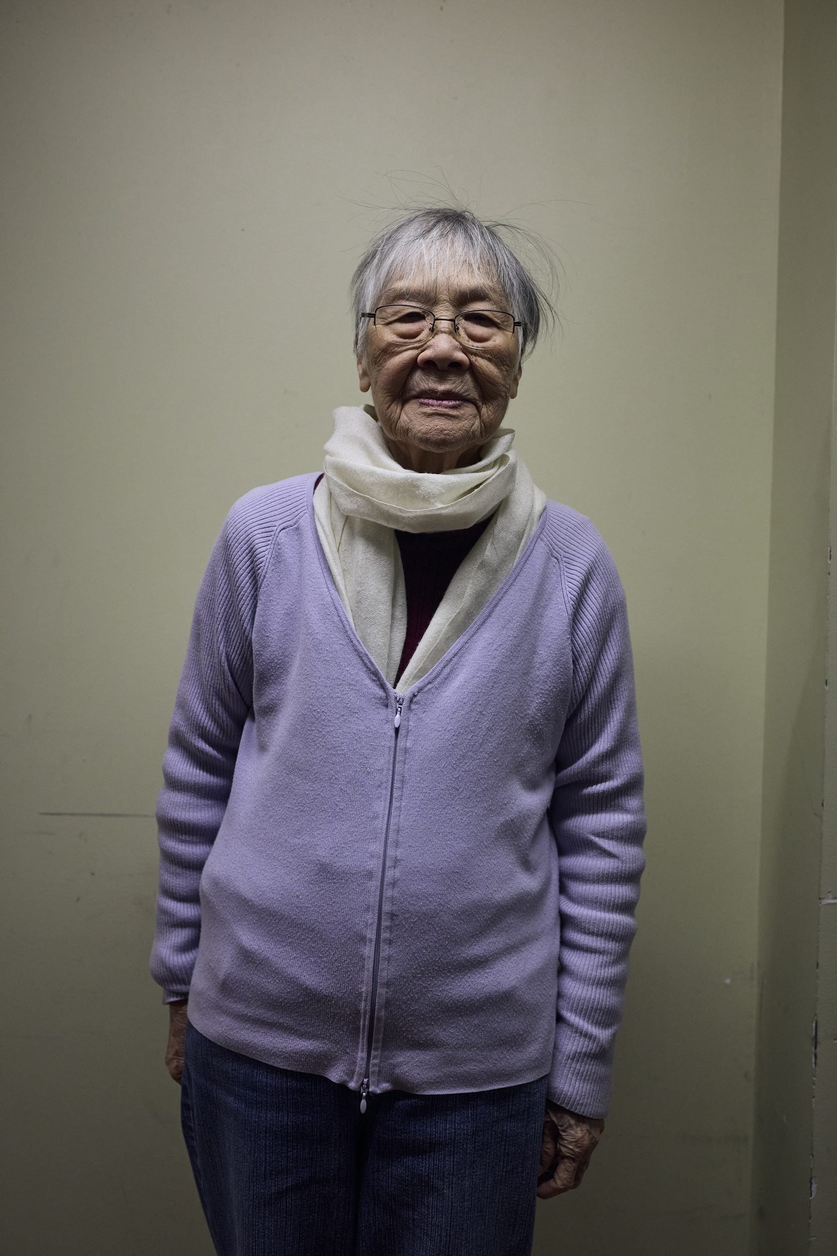 An elderly woman wearing glasses, a lavender cardigan, and a cream scarf stands against a plain wall.