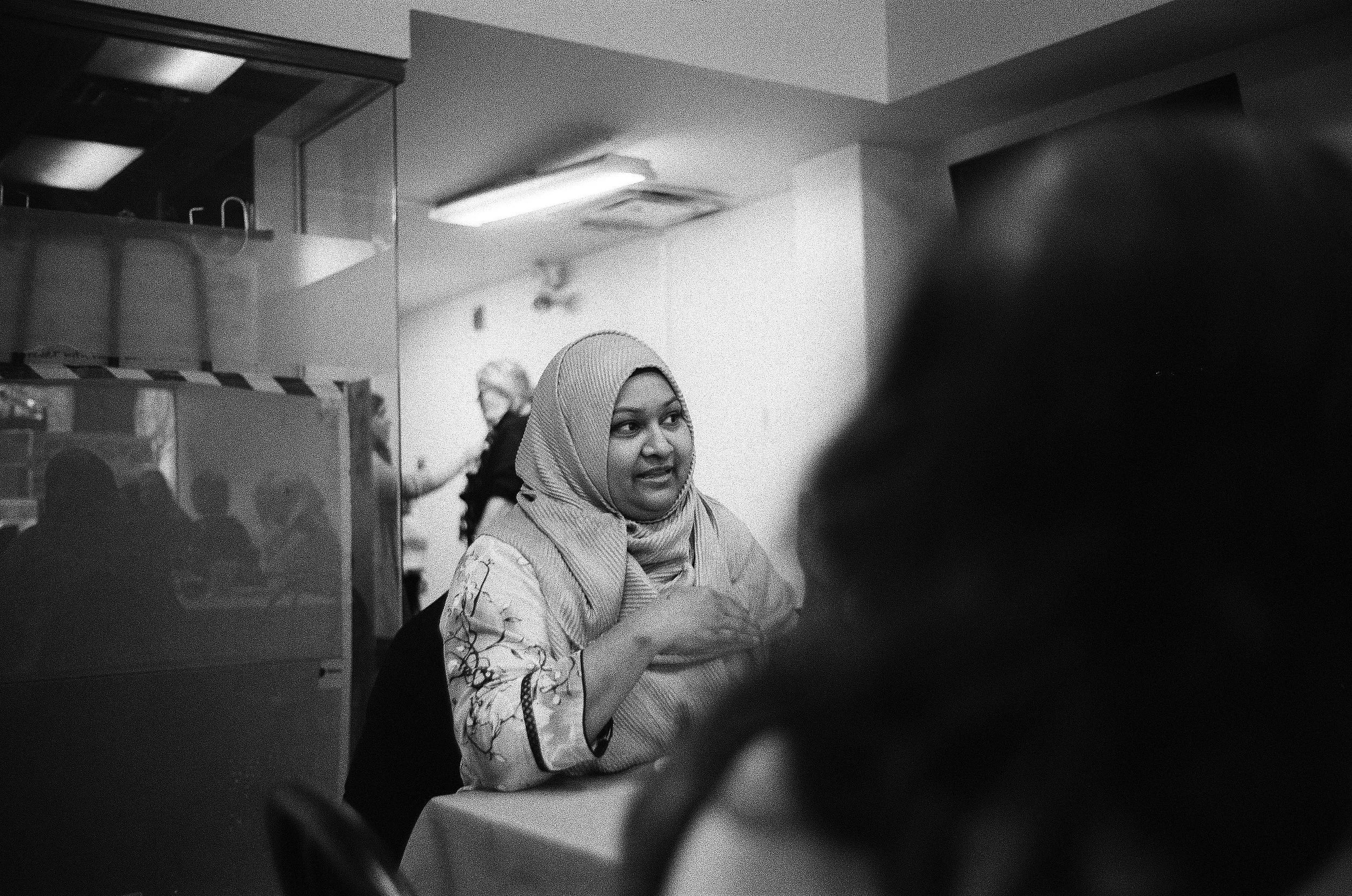 Woman wearing hijab and patterned clothing smiles in a crowded interior space lit by fluorescent lights.