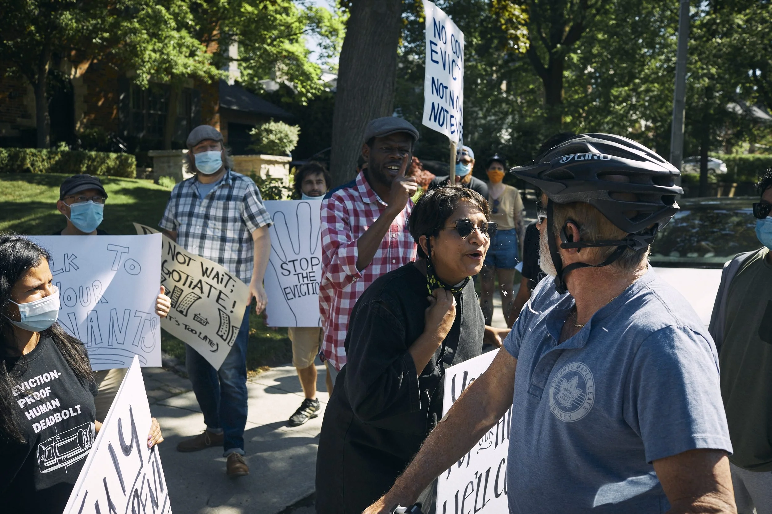 Protesters holding signs gather on a residential street with trees during a daytime demonstration.
