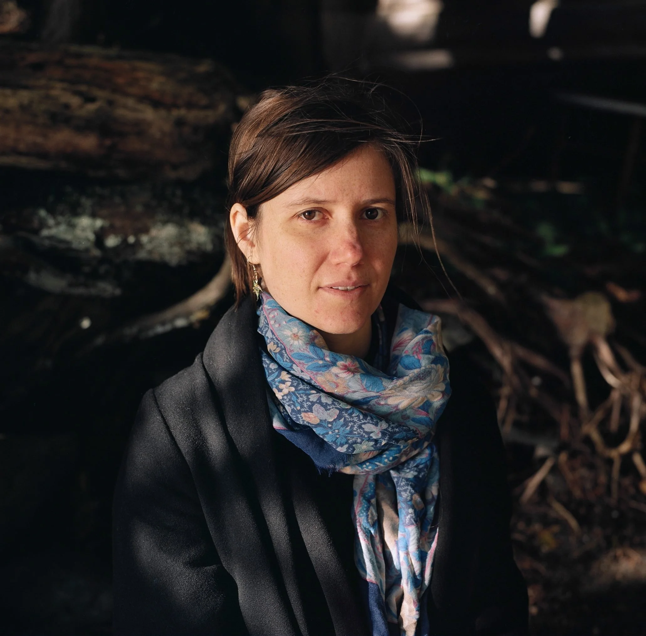 Woman in dark jacket and patterned scarf stands among fallen logs in wooded setting.