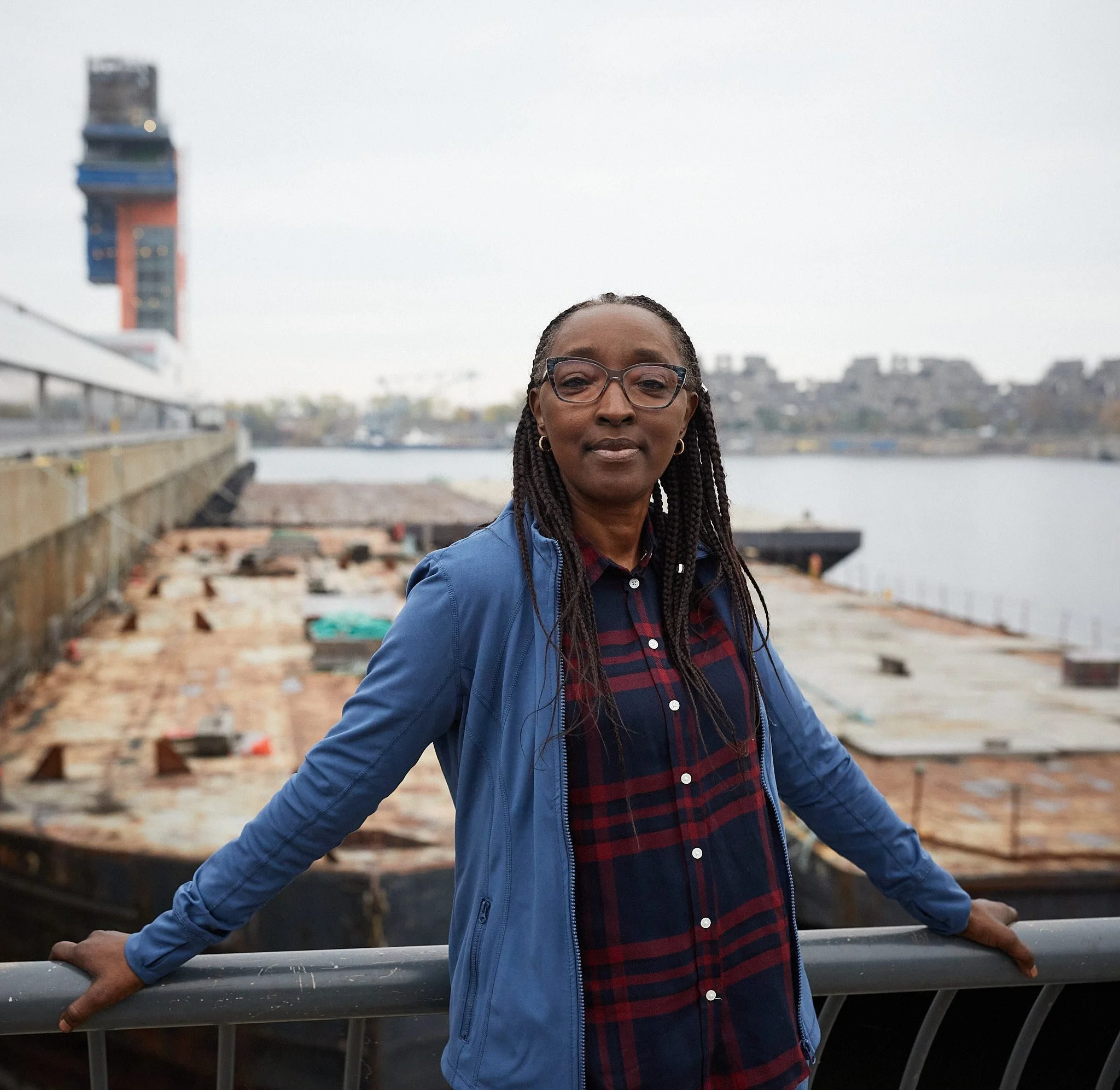 Woman in glasses and blue jacket poses at a waterfront with industrial docks and a tower in the background.