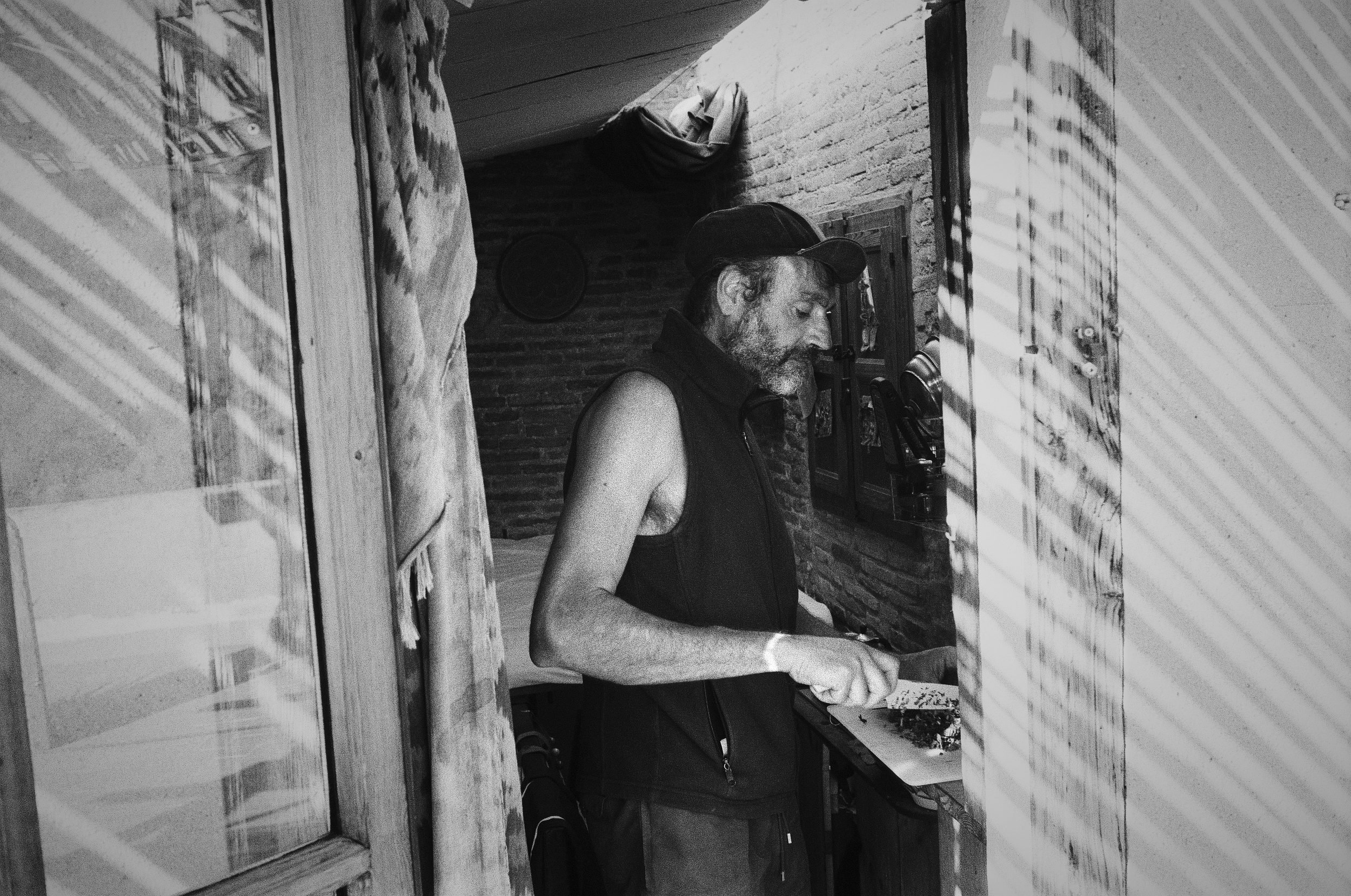A man in a baseball cap and sleeveless shirt works at a cluttered desk in a narrow brick room.