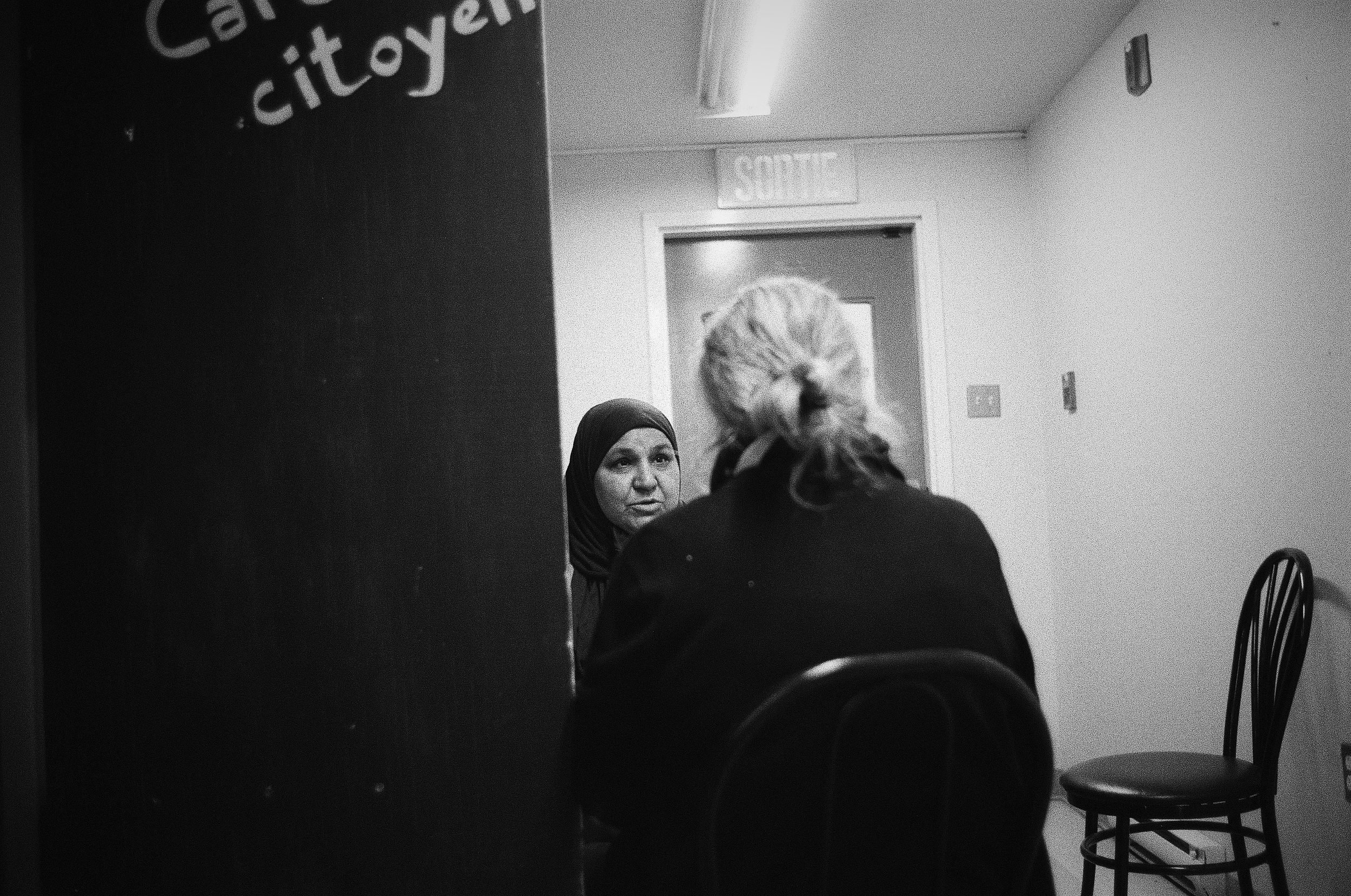 Two women converse in a narrow hallway with a chair and doorway visible.
