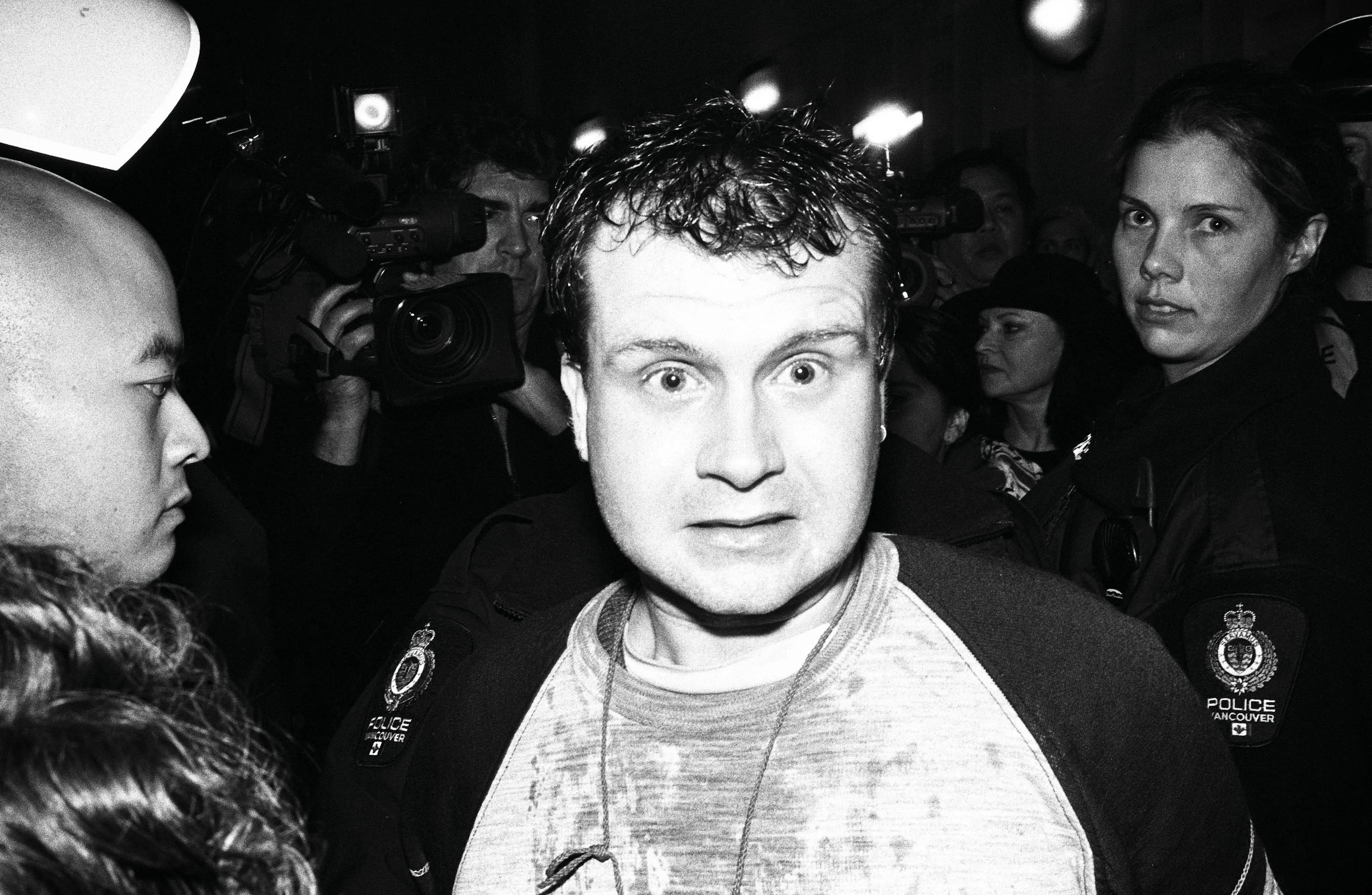 Man in gray shirt surrounded by police officers and photographers at night.