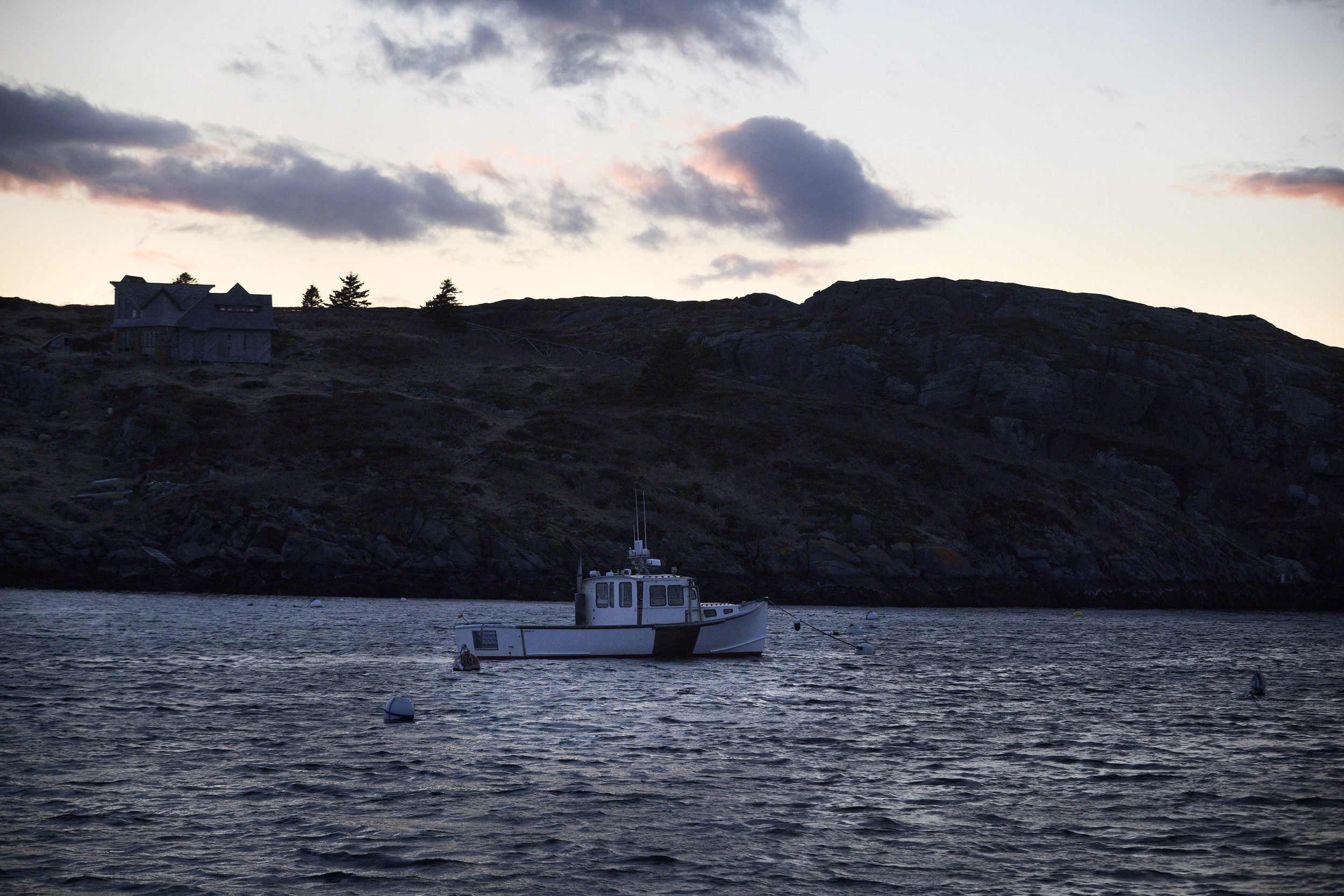A white fishing boat anchors in calm water near rocky cliffs with a house silhouetted on the hillside at dusk.