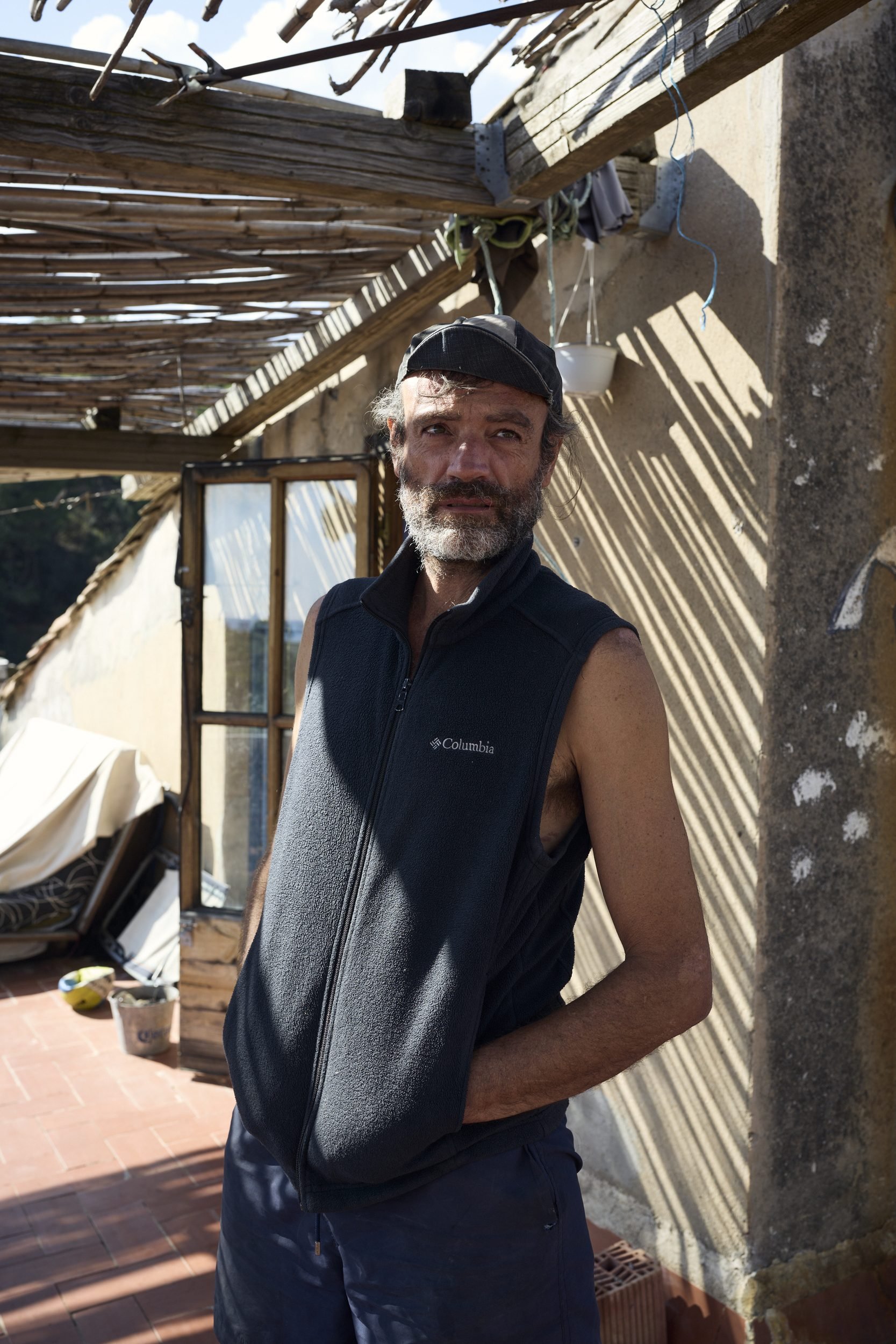 A man with a beard wearing a Columbia vest and baseball cap stands on a sunny rooftop with wooden structures and concrete walls.