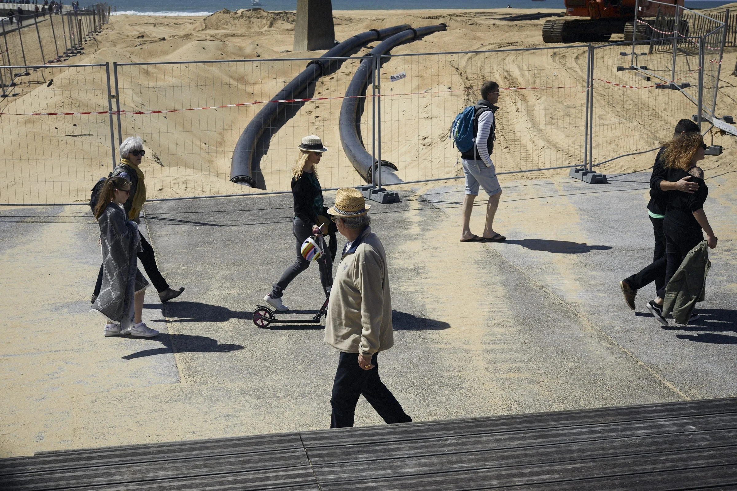 Several people walk and ride scooters on a wooden boardwalk at a beach enclosed by metal fencing.