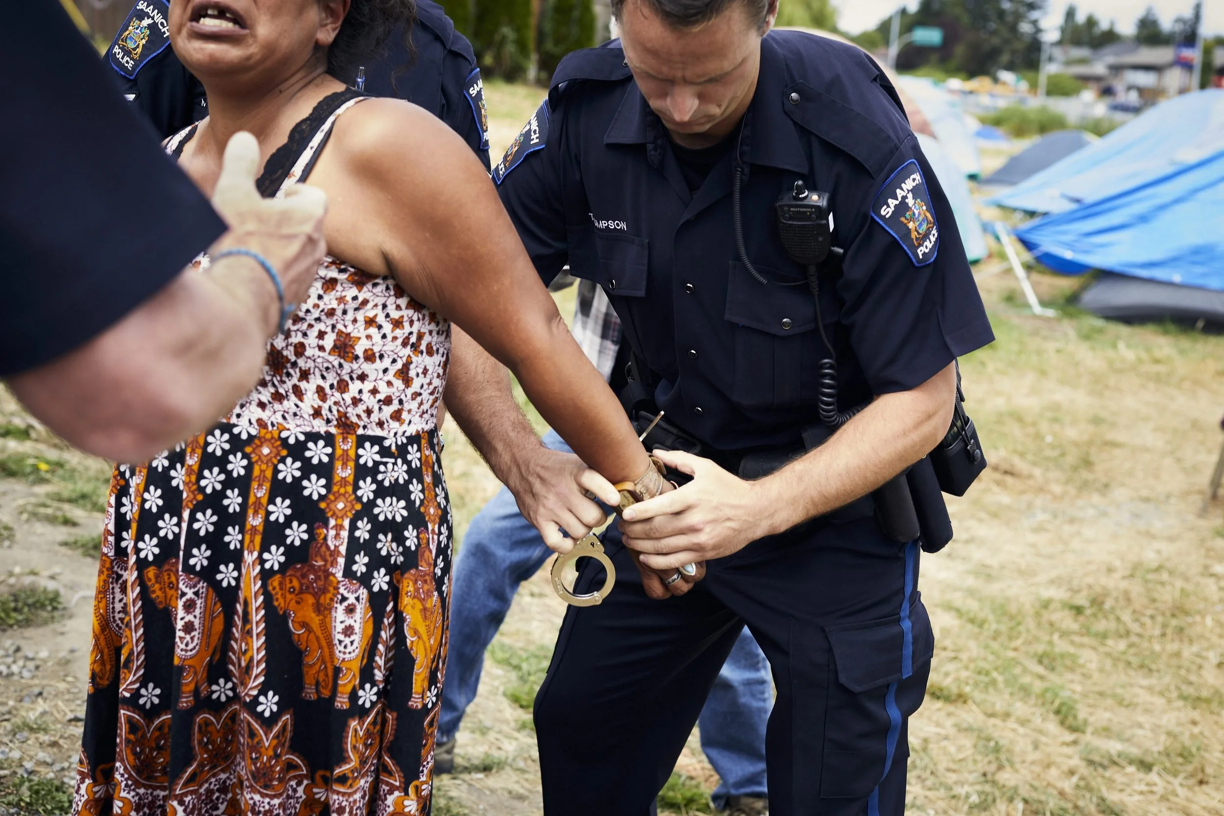 Police officer handcuffs a person at an outdoor event with blue tarps visible in the background.