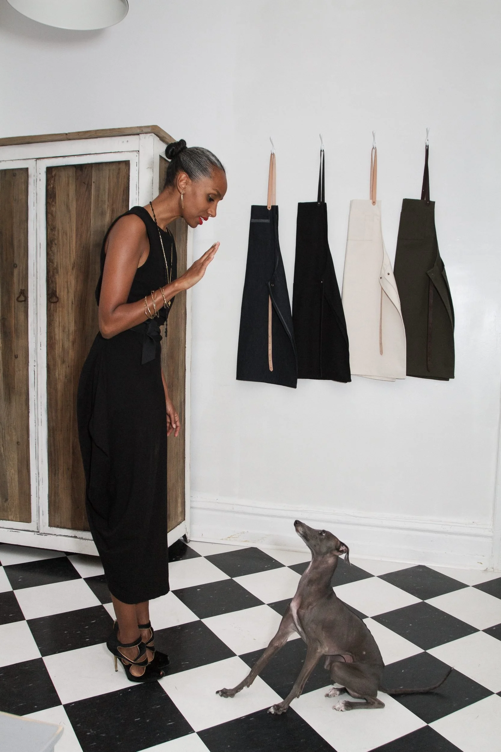 Woman in black dress gestures toward hanging fabric swatches while a dog sits on a checkered floor beside her.