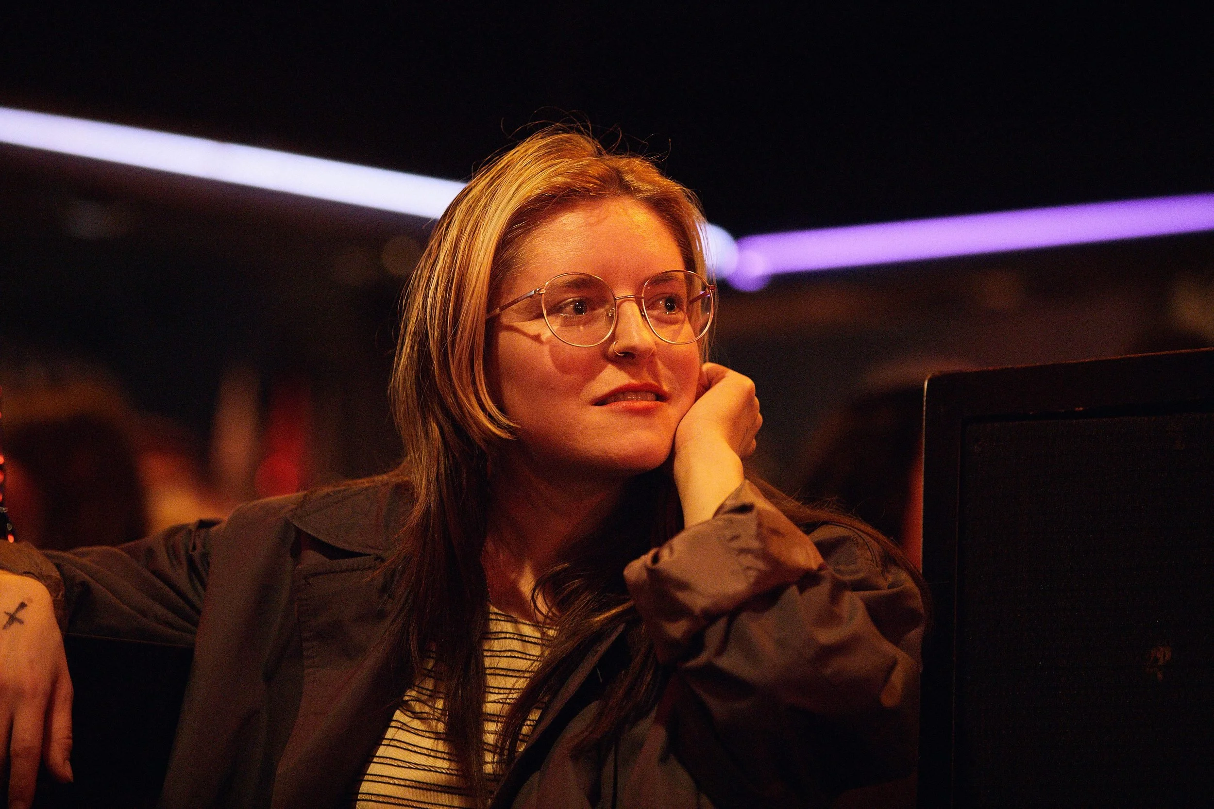 Woman wearing glasses and dark jacket rests her chin on her hand in warm stage lighting.