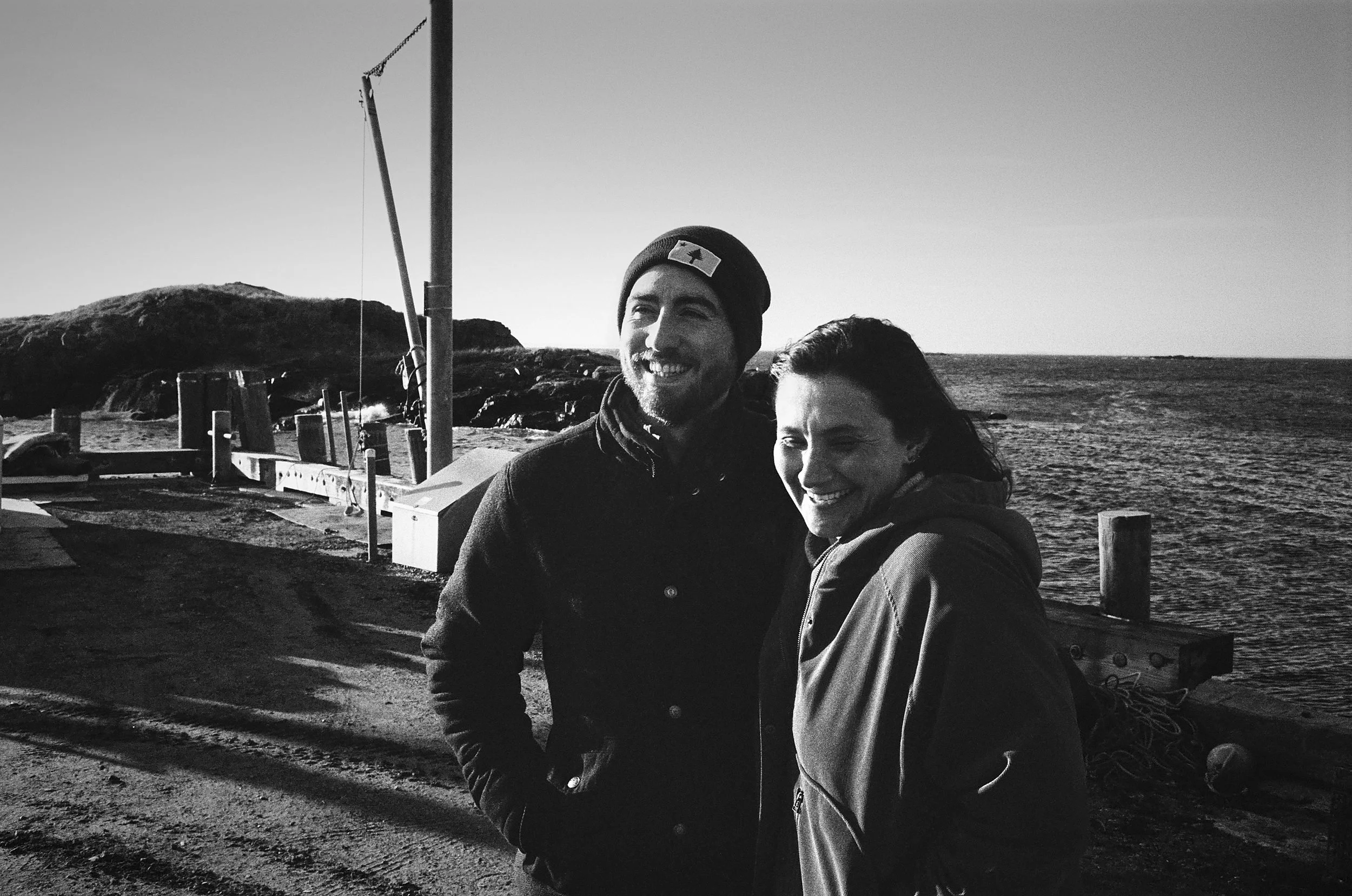 Two people smile at the camera on a beach with boats and rocky coastline visible behind them.