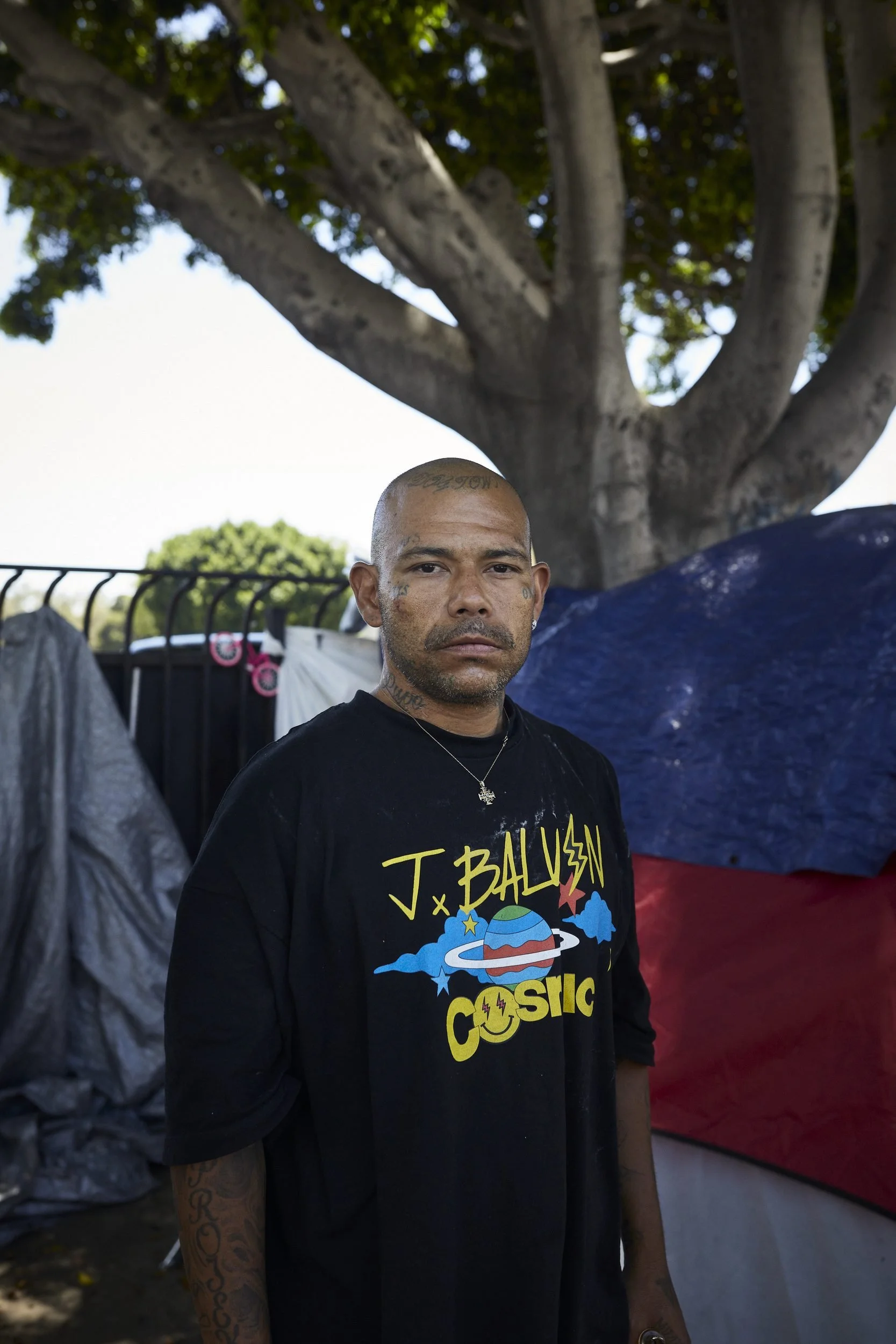 A man in a black graphic t-shirt stands beneath a large tree, with clotheslines and colored fabric visible behind him.
