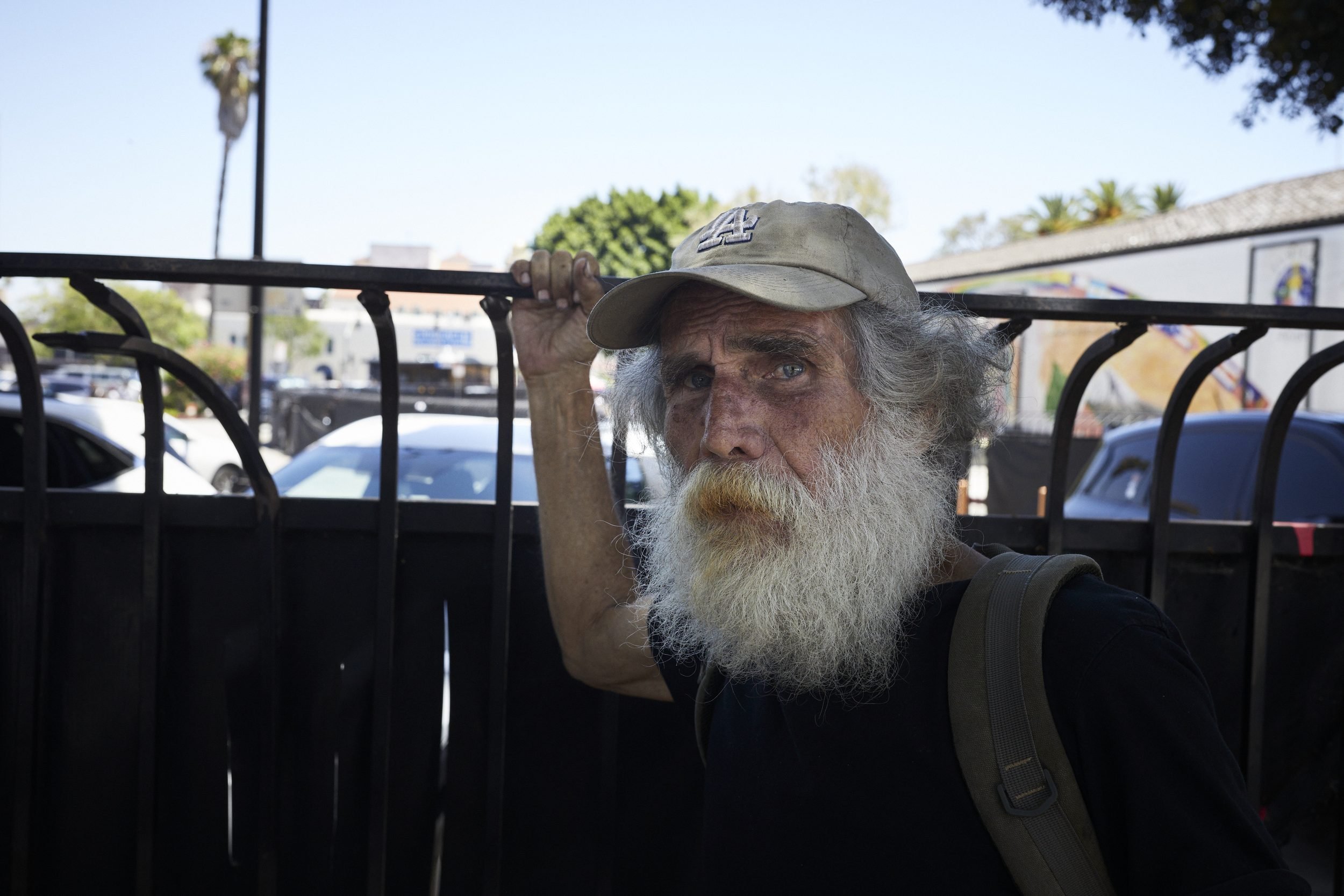 An older man with a long white beard wearing a cap holds onto a black metal railing in an urban setting.