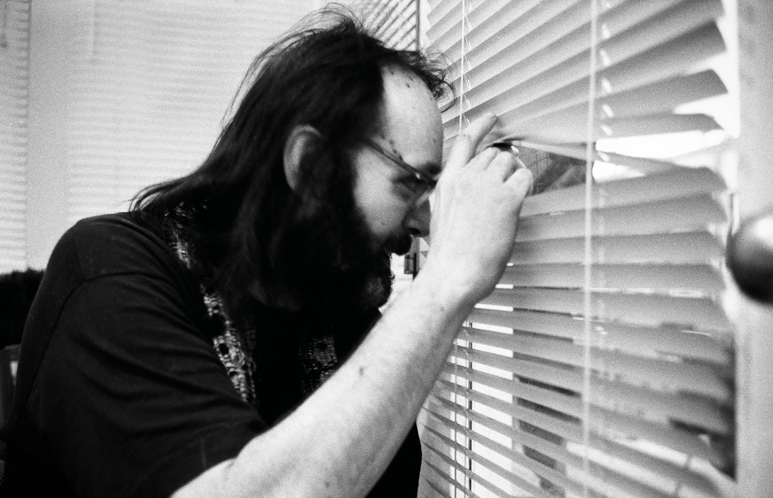 Man with long hair and glasses peers through window blinds in black and white photograph.