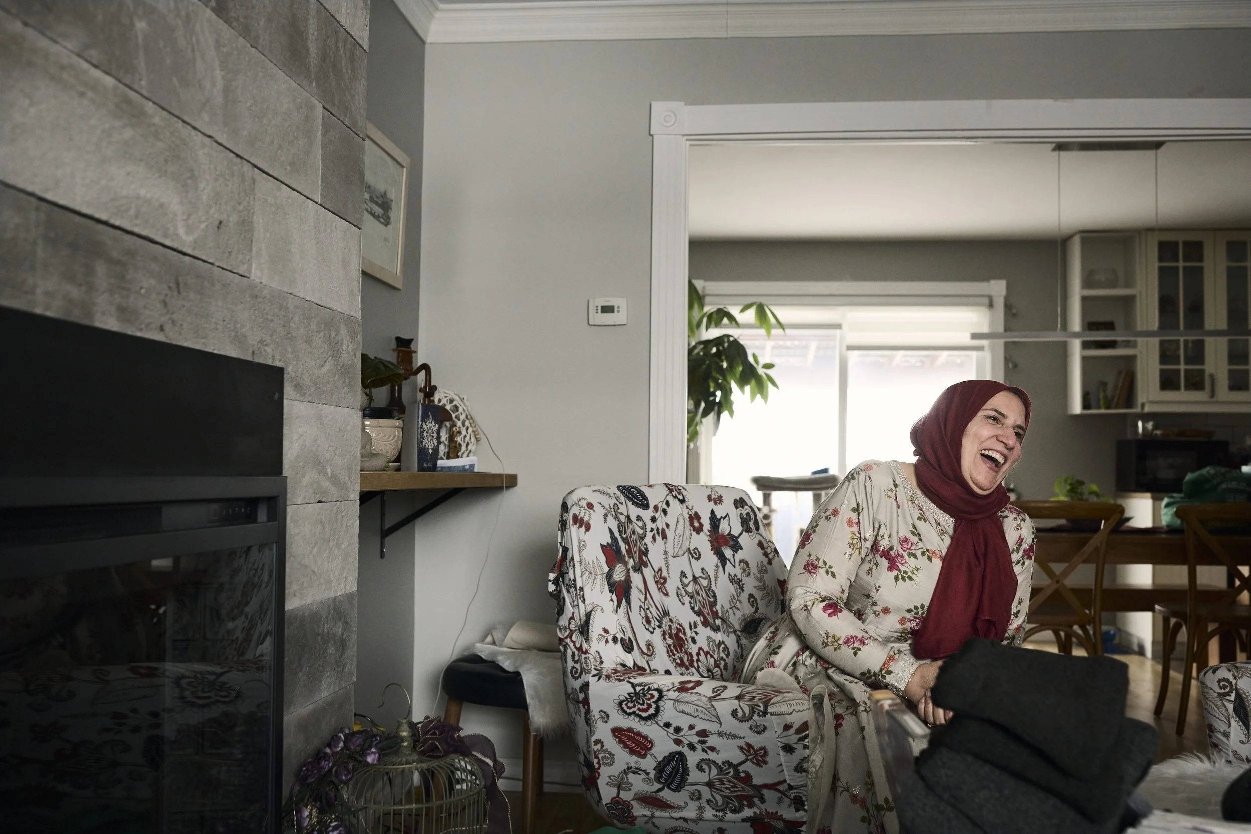 Woman in a floral chair laughs while sitting in a modern living room with a fireplace.
