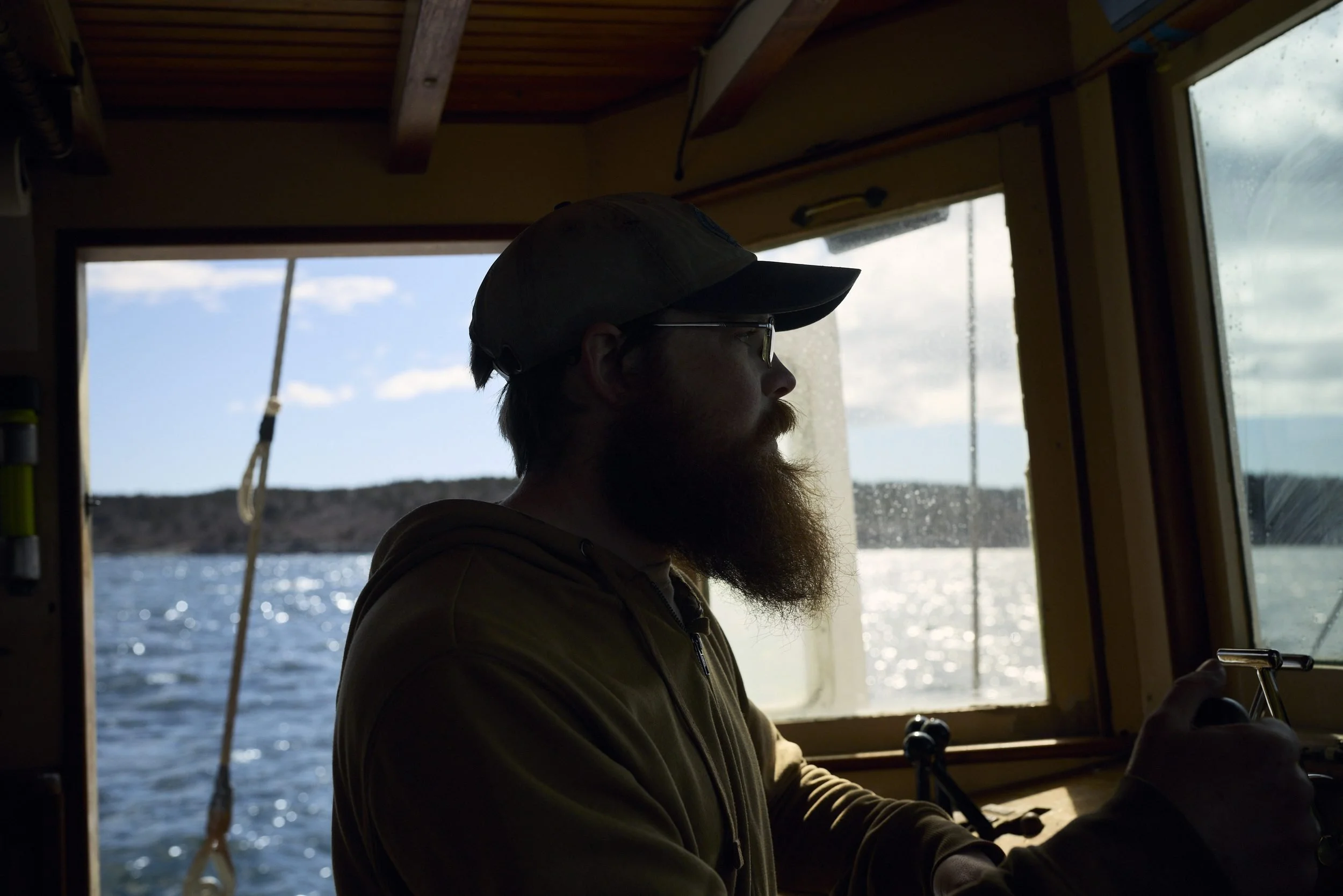 Man with a beard at the helm of a boat, looking out at water and shoreline through cabin windows.