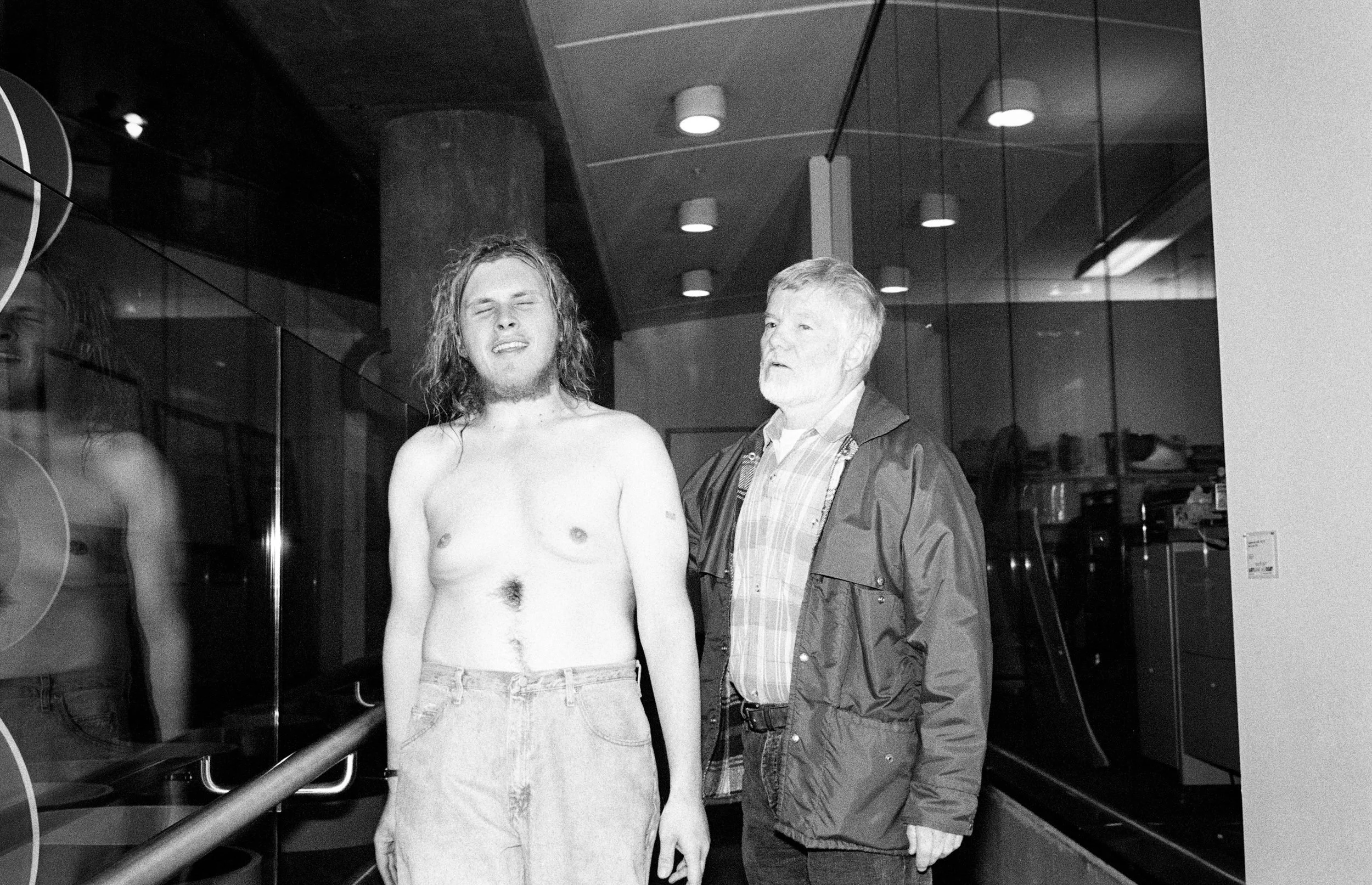 Two men stand in a modern indoor corridor with recessed lighting and glass panels.