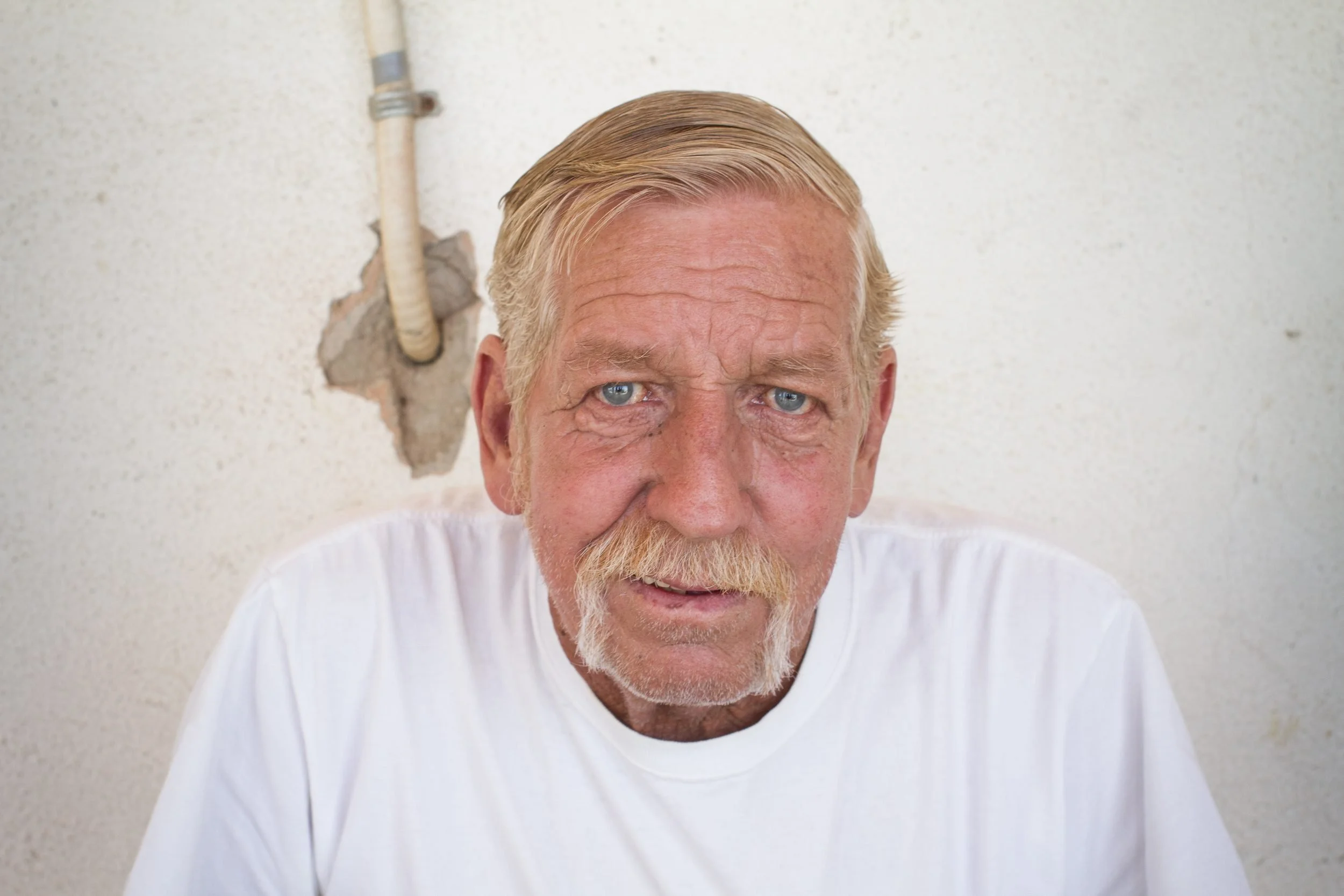 Older man with blonde hair and mustache wearing white t-shirt against plain wall with rope visible in background.