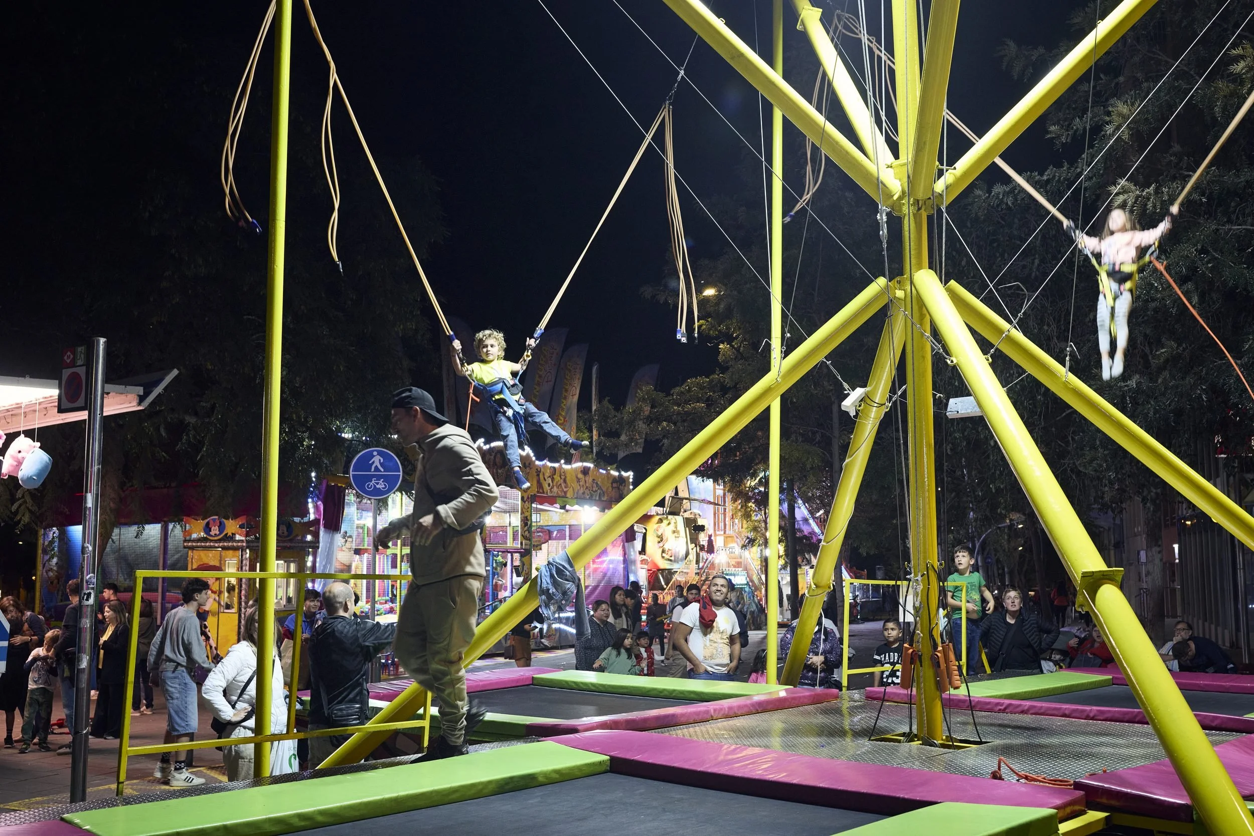 Acrobats perform on a brightly lit yellow trapeze structure at a nighttime outdoor festival or carnival.