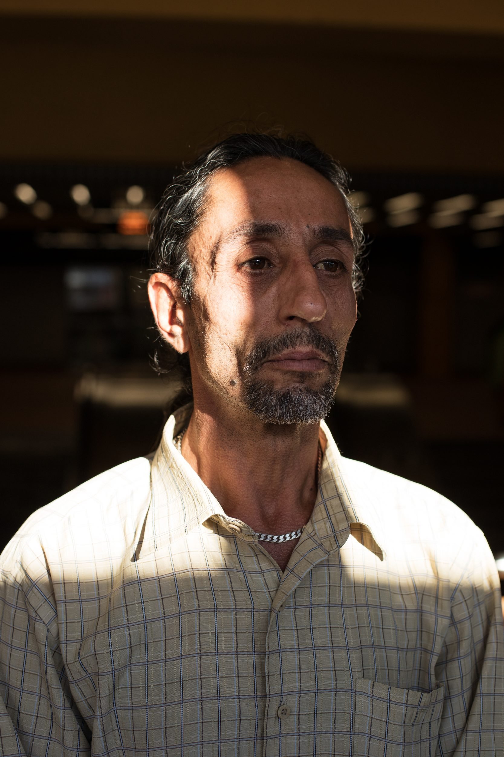 Portrait of a bearded man in a light plaid shirt against a dark interior background.