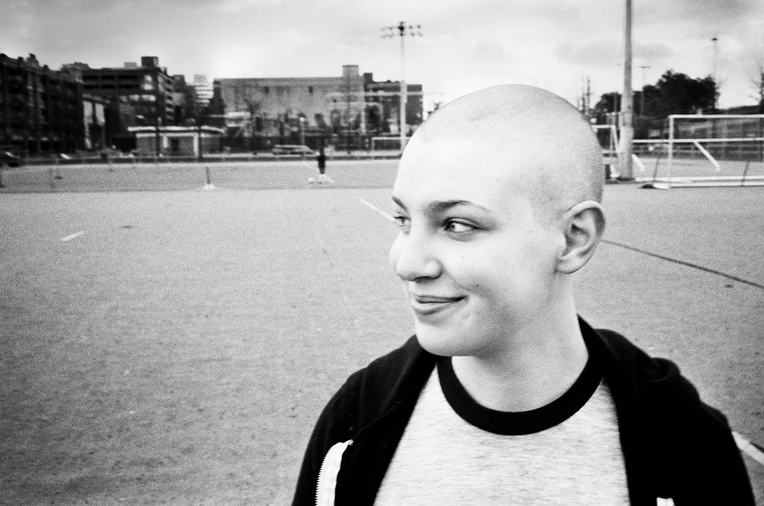 Person with shaved head smiling at an outdoor sports court with buildings in the background.