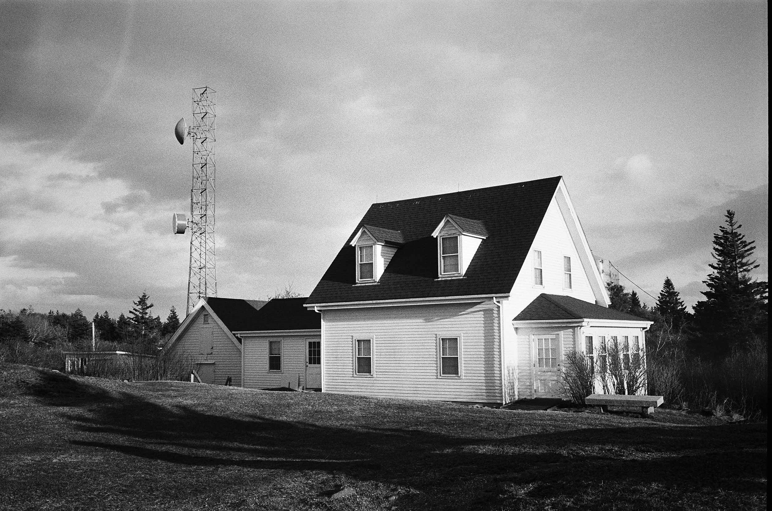 White farmhouse with dormer windows beside a tall communication tower under cloudy sky.