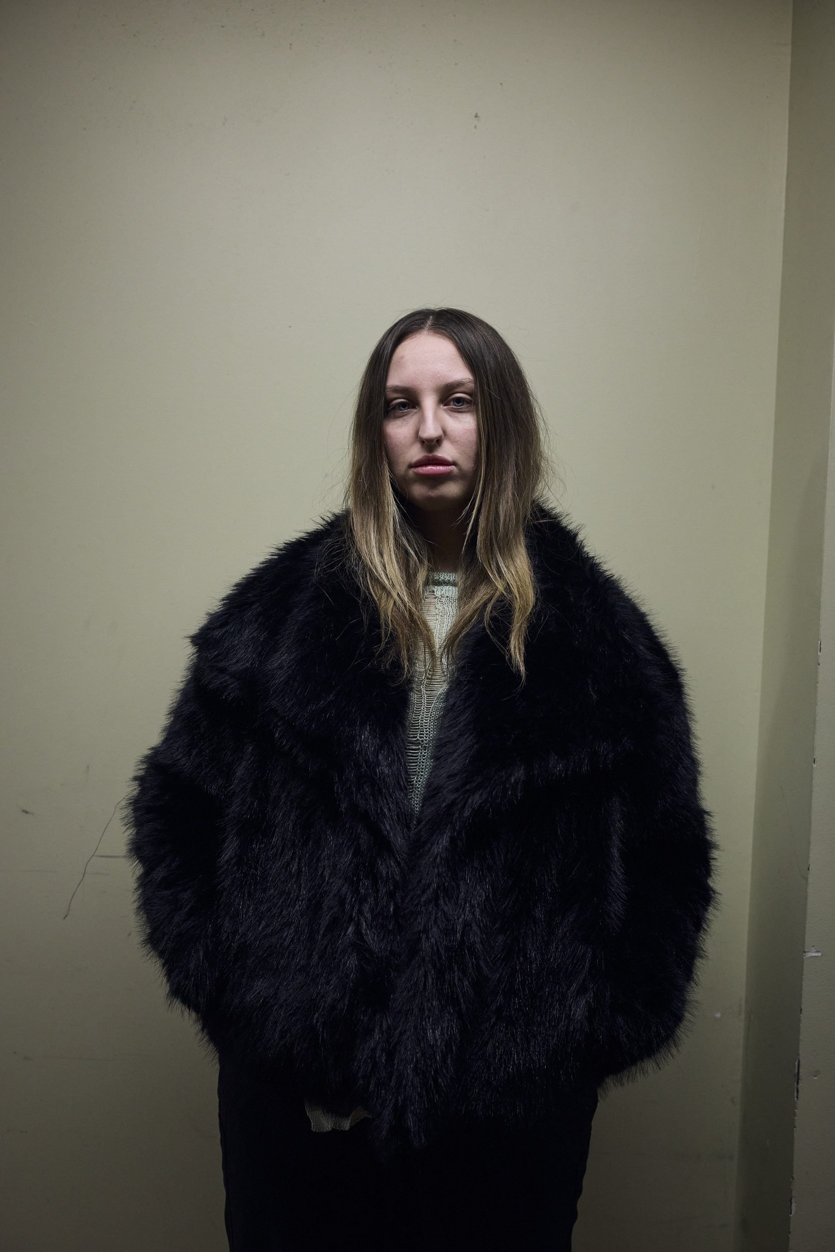 Woman in black fur stole and gray sweater stands against pale wall, looking at camera.