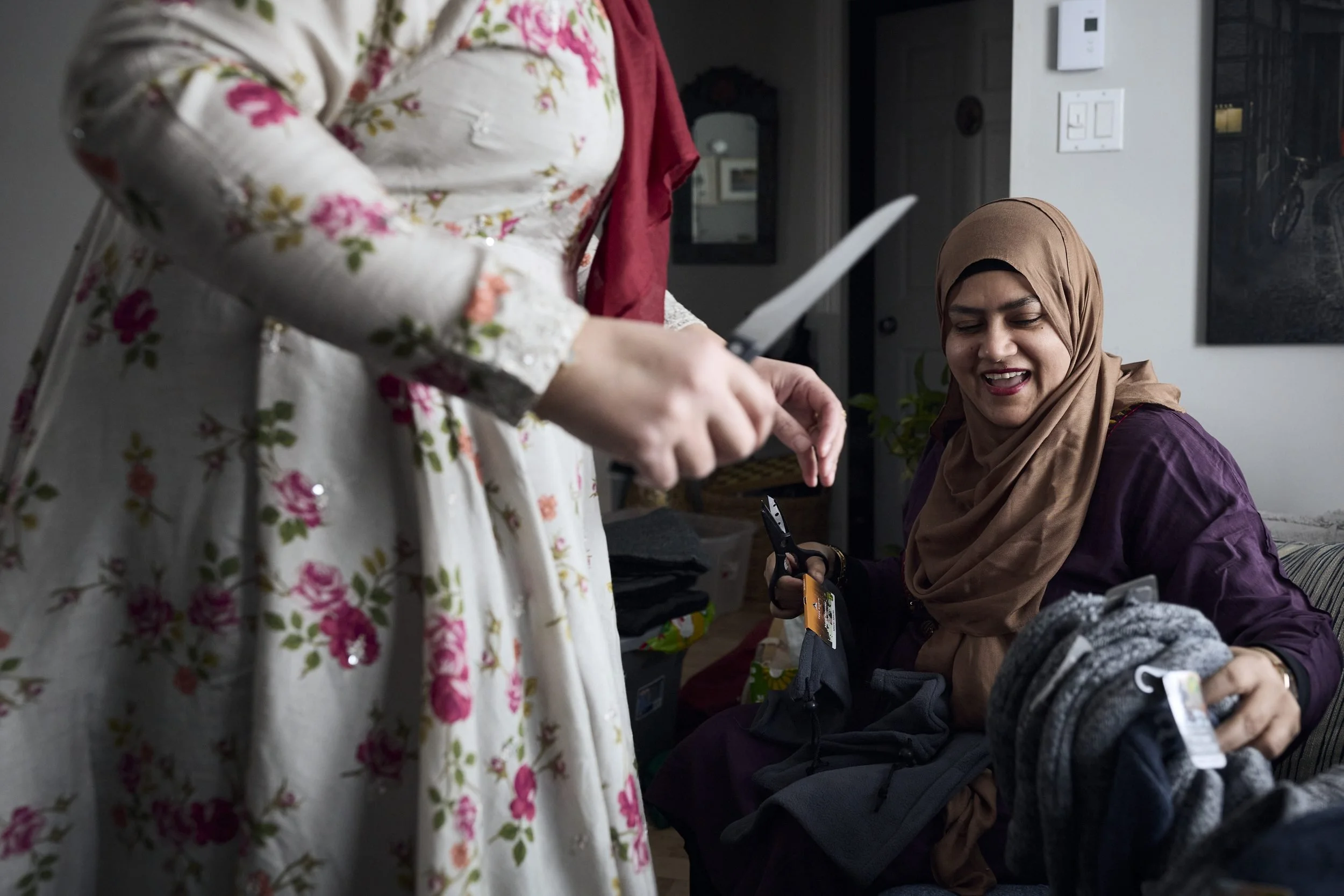 Two women sit together indoors, one wearing a beige hijab and purple top smiling while holding clothing items.