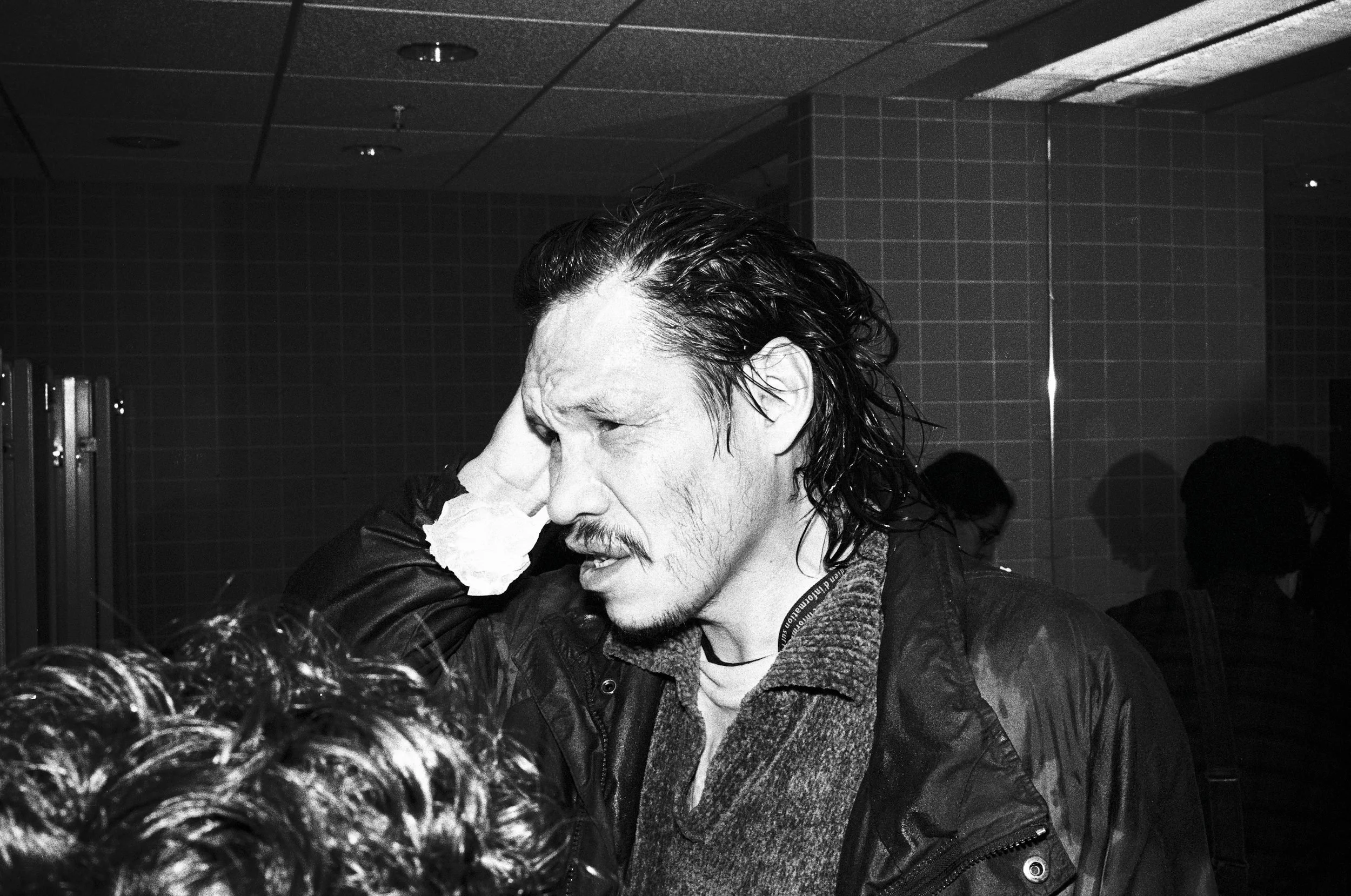 Man with long dark hair and mustache holding tissue to his face in an indoor setting.