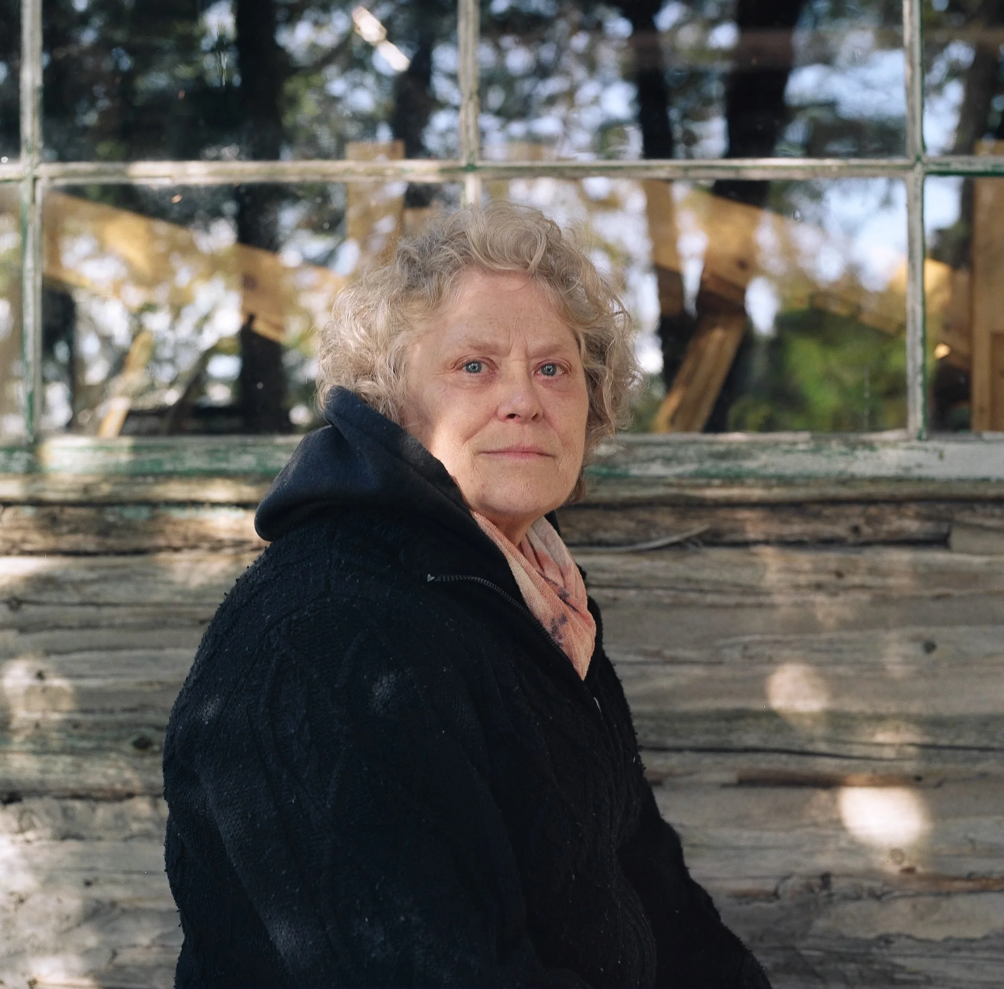 An older woman with gray curled hair wearing a black jacket stands on a wooden deck facing the camera, with a glass-paneled structure behind her.