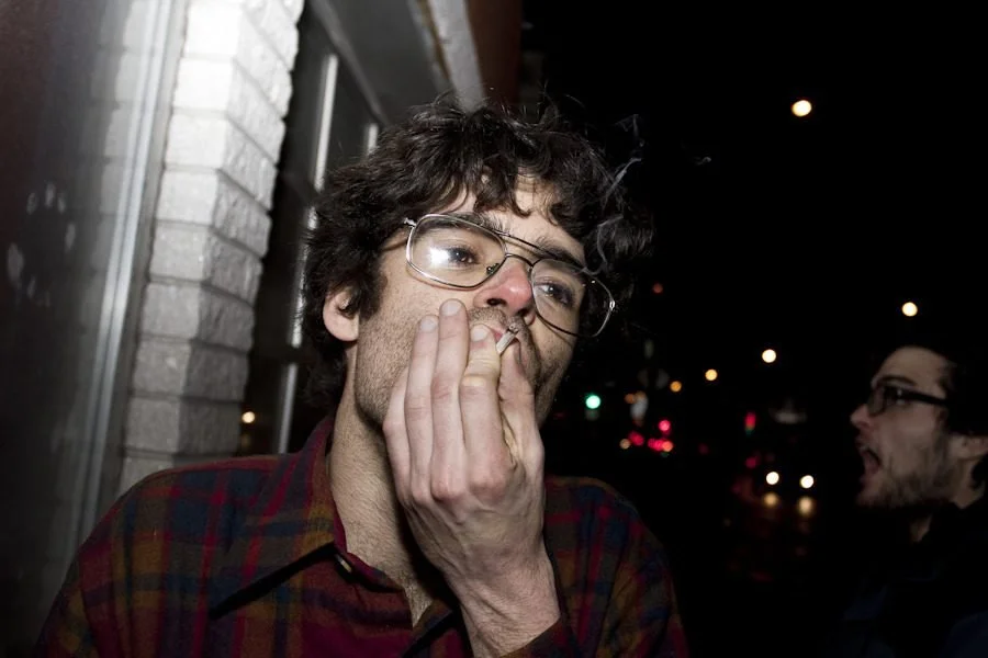 Man wearing glasses and plaid shirt covers his mouth while standing on a nighttime city street.