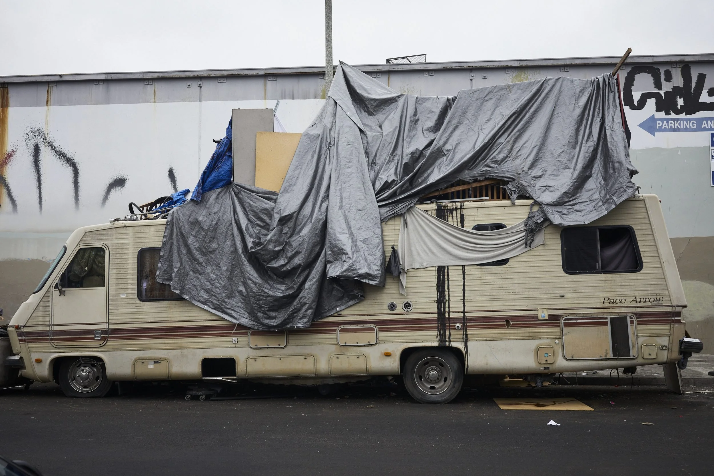 A weathered RV covered with dark tarps is parked against an industrial building with graffiti.