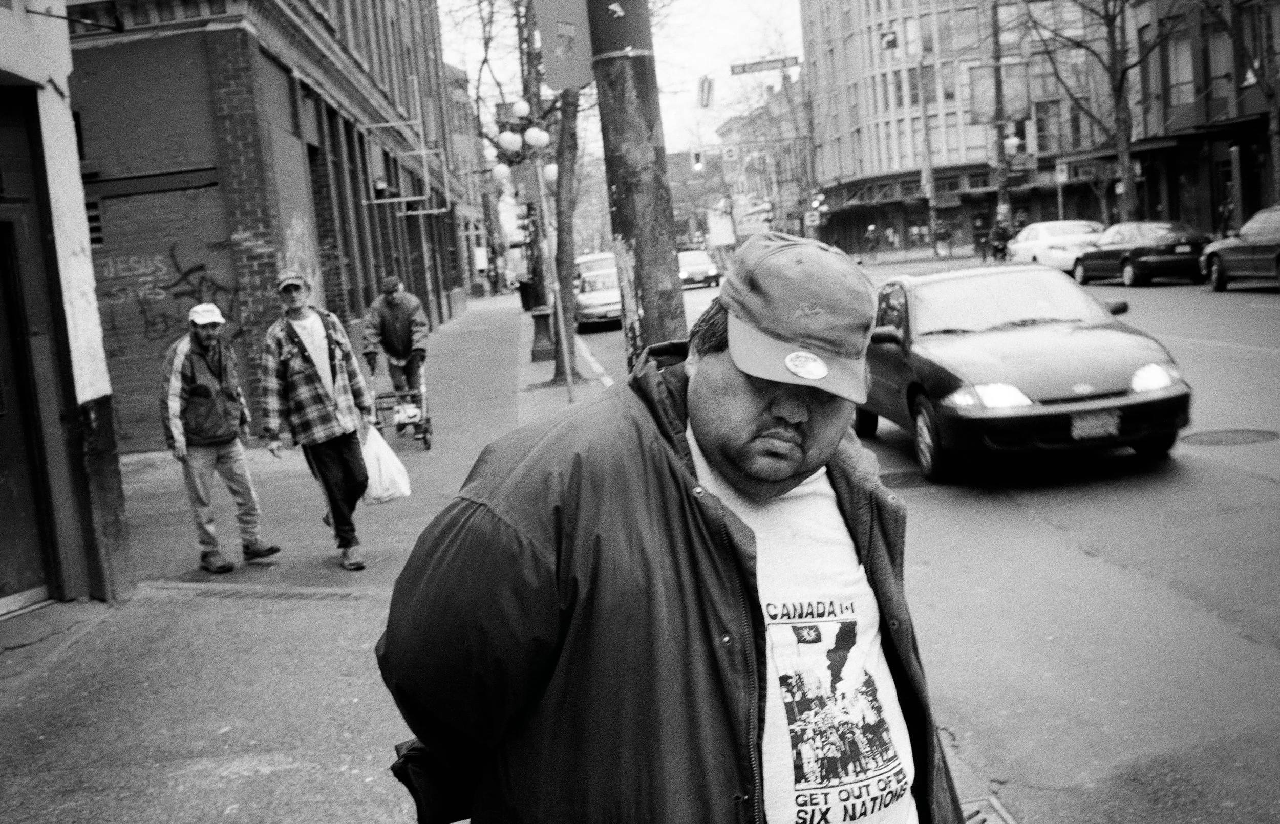 Man wearing Canada-themed shirt and cap stands on urban street with buildings and parked cars behind him.
