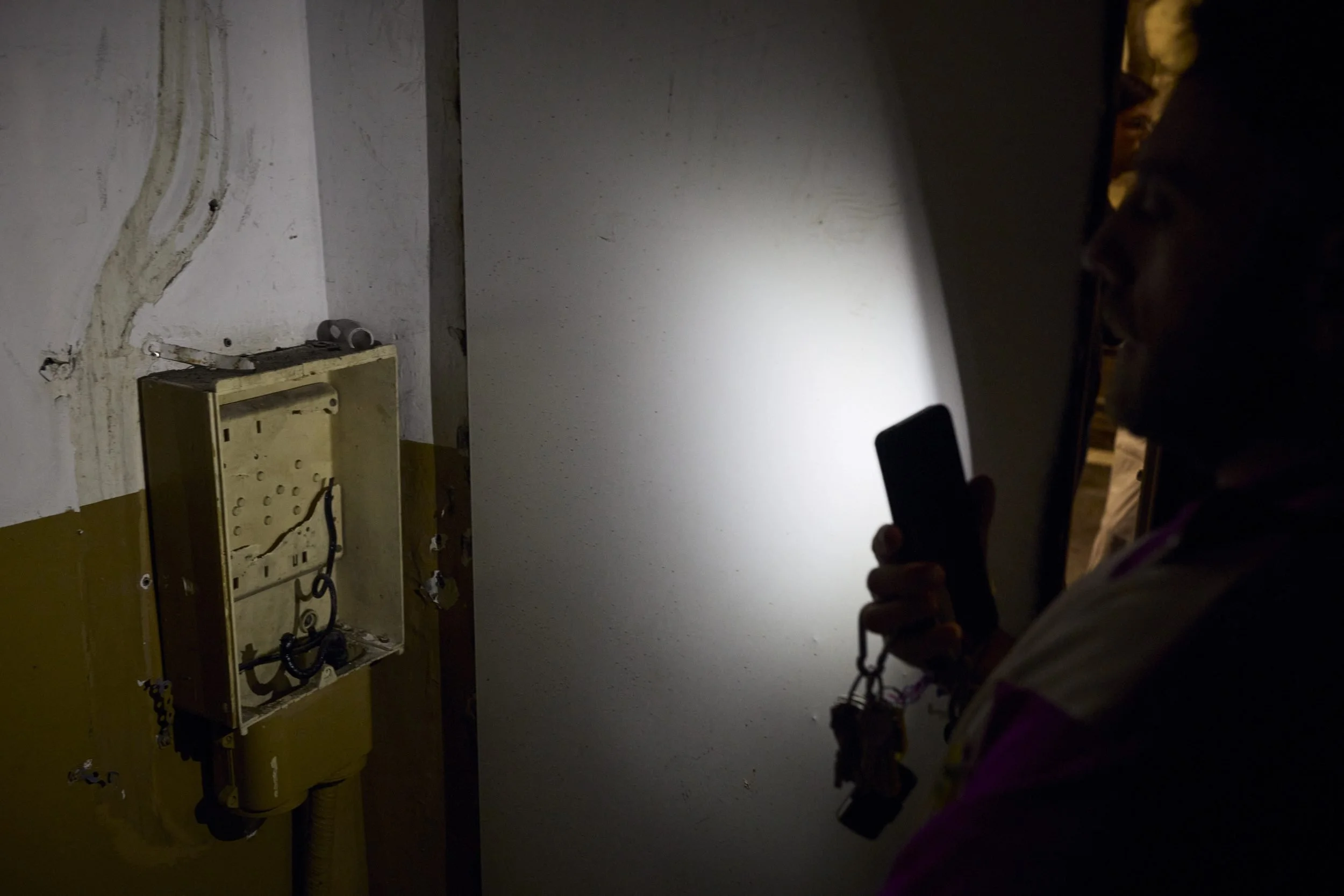 A person holding a smartphone photographs an open electrical panel on a yellow-painted wall.