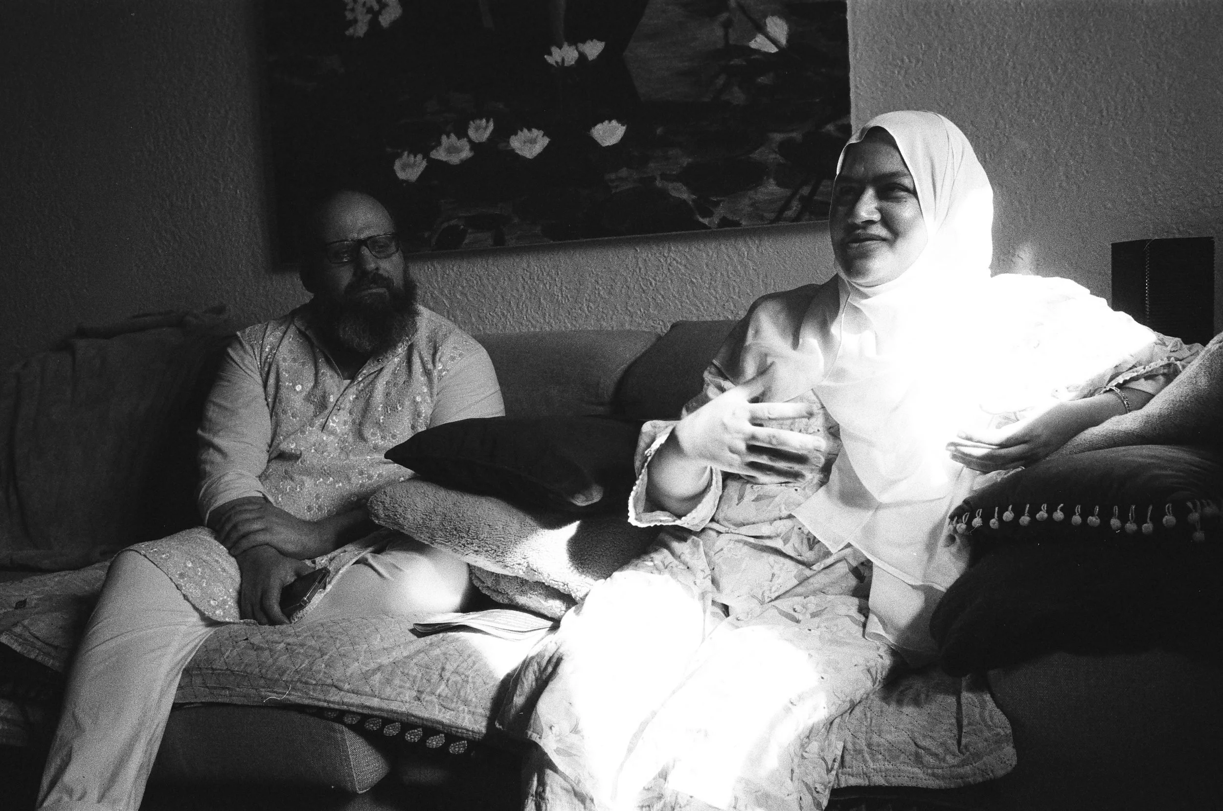 Two people sit on a couch with papers and documents, one wearing glasses and a beard, the other wearing a white head covering.