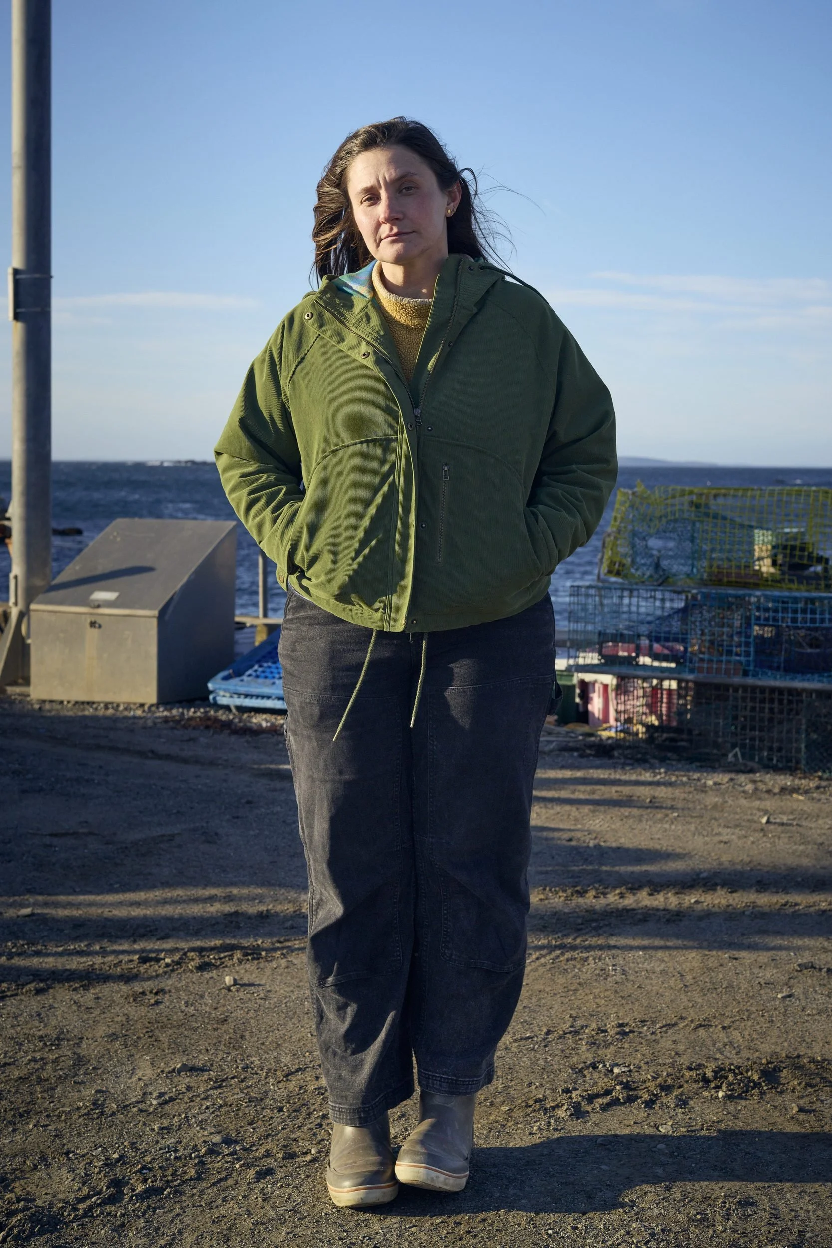Woman in olive jacket standing on gravel near water and stacked fishing traps.