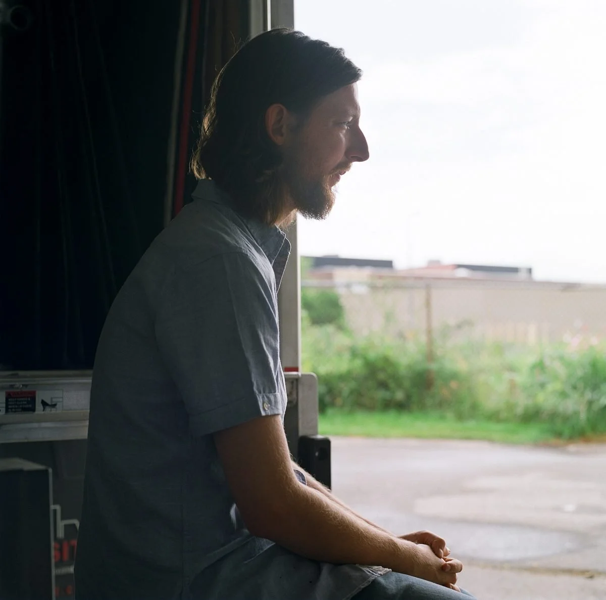 Man with long hair and beard gazes out a window toward agricultural fields.