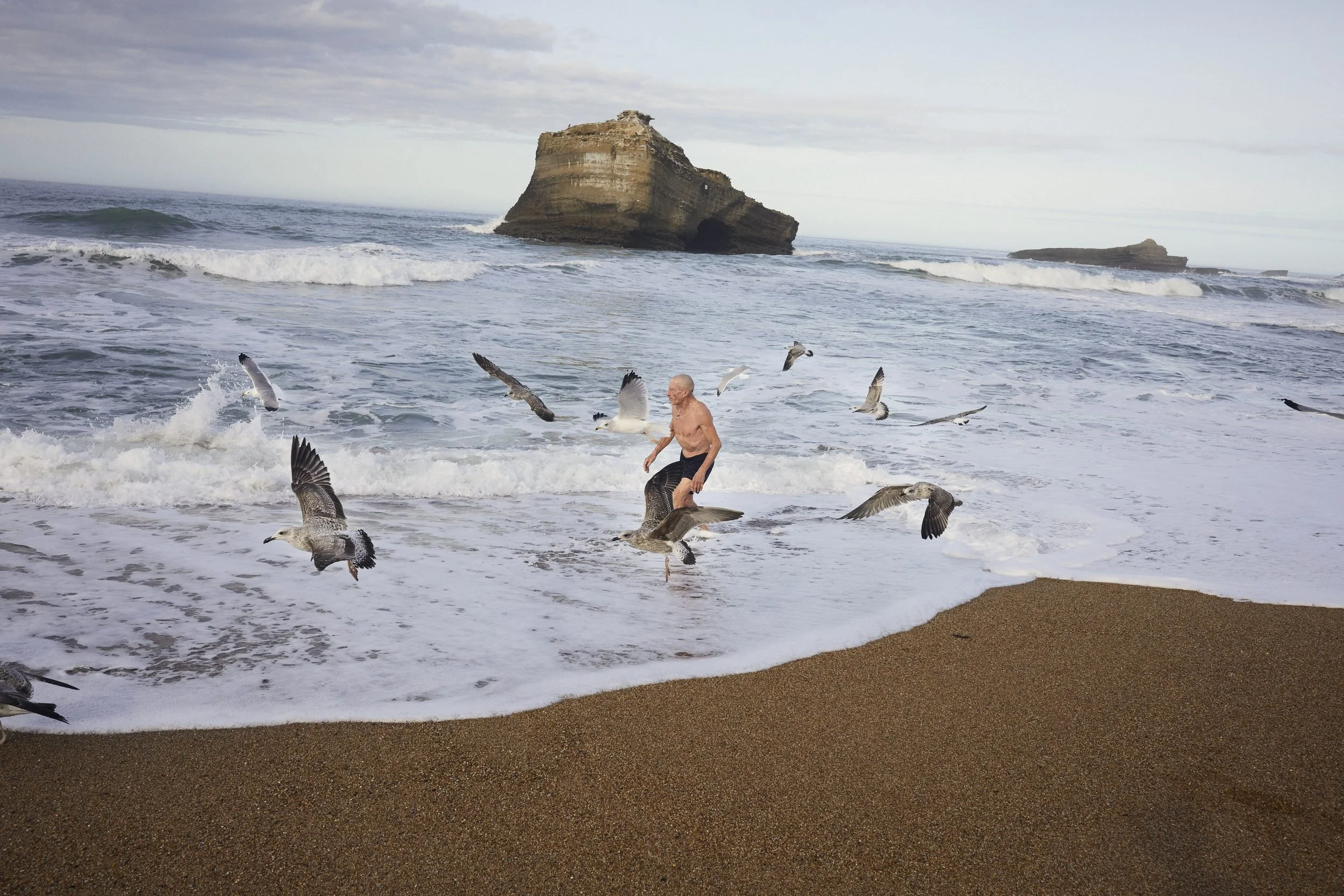 A shirtless man wades in the ocean surrounded by flying birds, with a large rock formation visible in the background.
