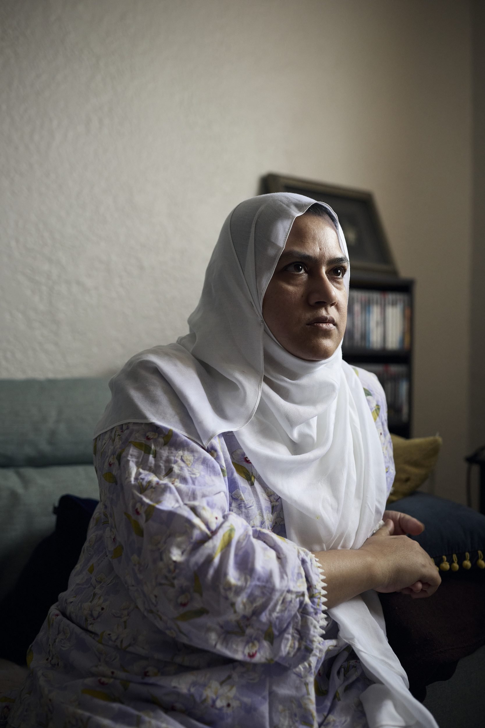 Woman wearing a white hijab and patterned garment sitting on a couch, looking to the side.