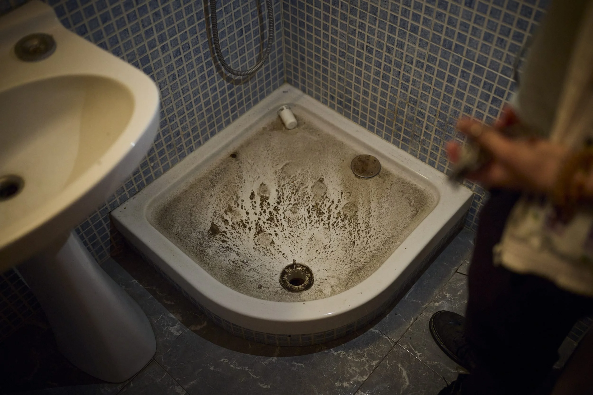 A neglected bathroom sink filled with sand or debris sits next to a toilet in a tiled room.