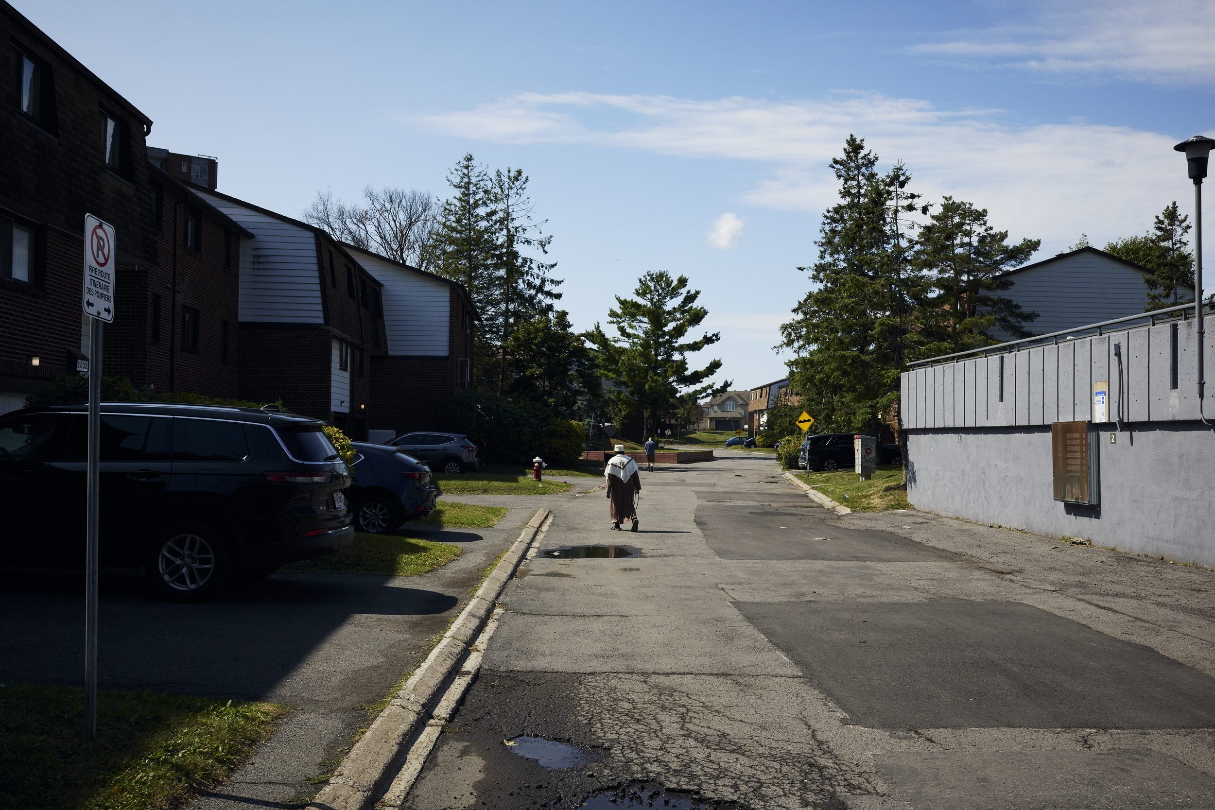 A person walks down a residential street lined with parked cars and modest apartment buildings on a sunny day.