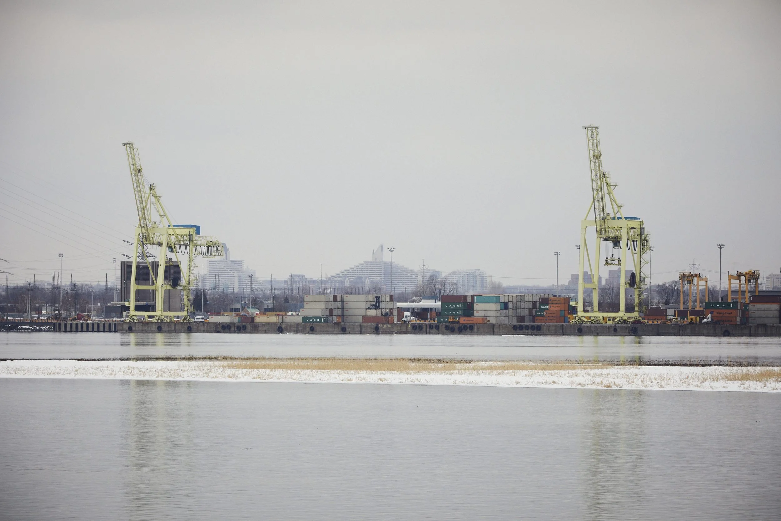 Two large yellow shipping cranes stand on a frozen riverbank lined with stacked cargo containers and industrial buildings.
